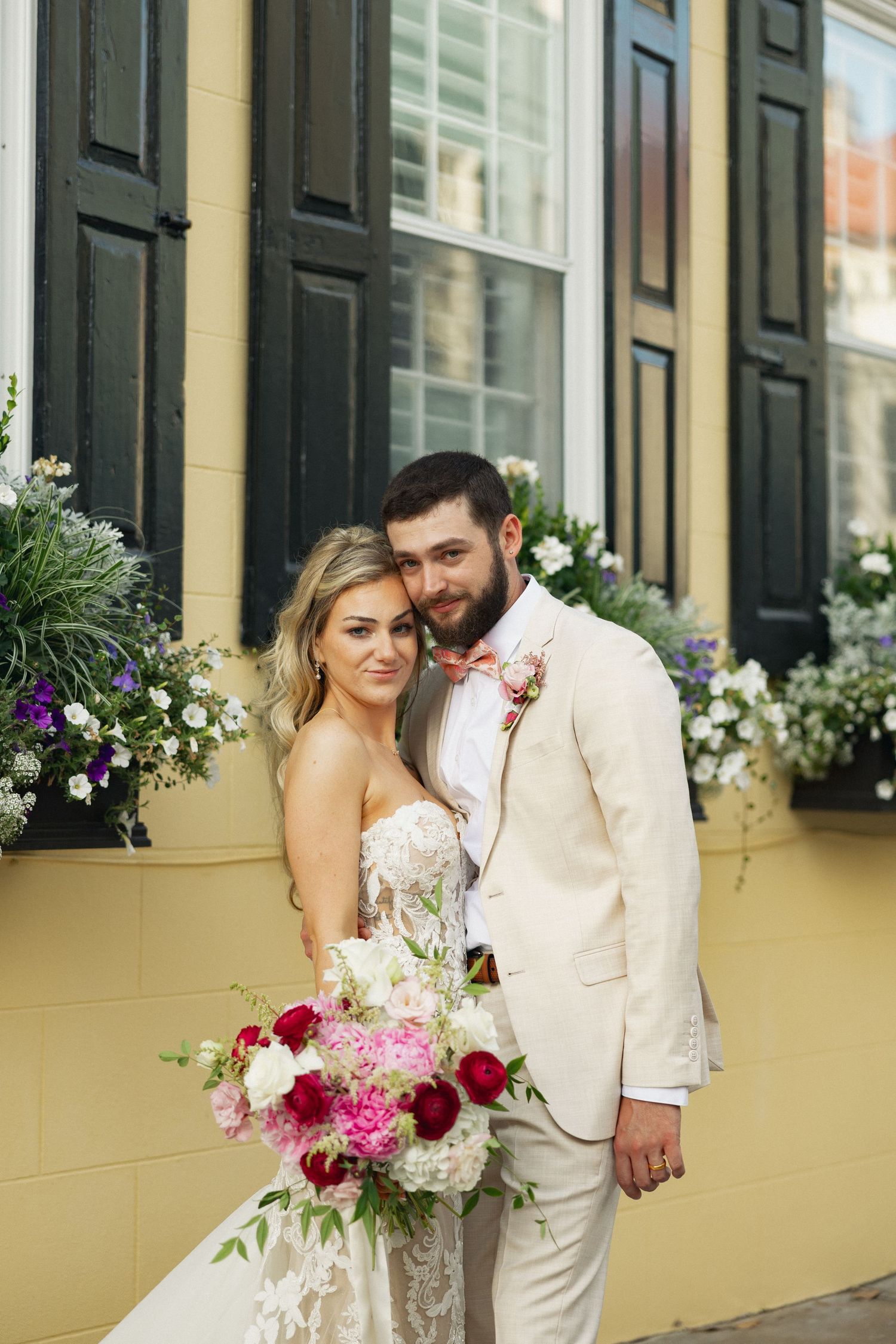 Groom in cream suit embraces bride holding vibrant pink and white bouquet beside yellow building with black shutters.