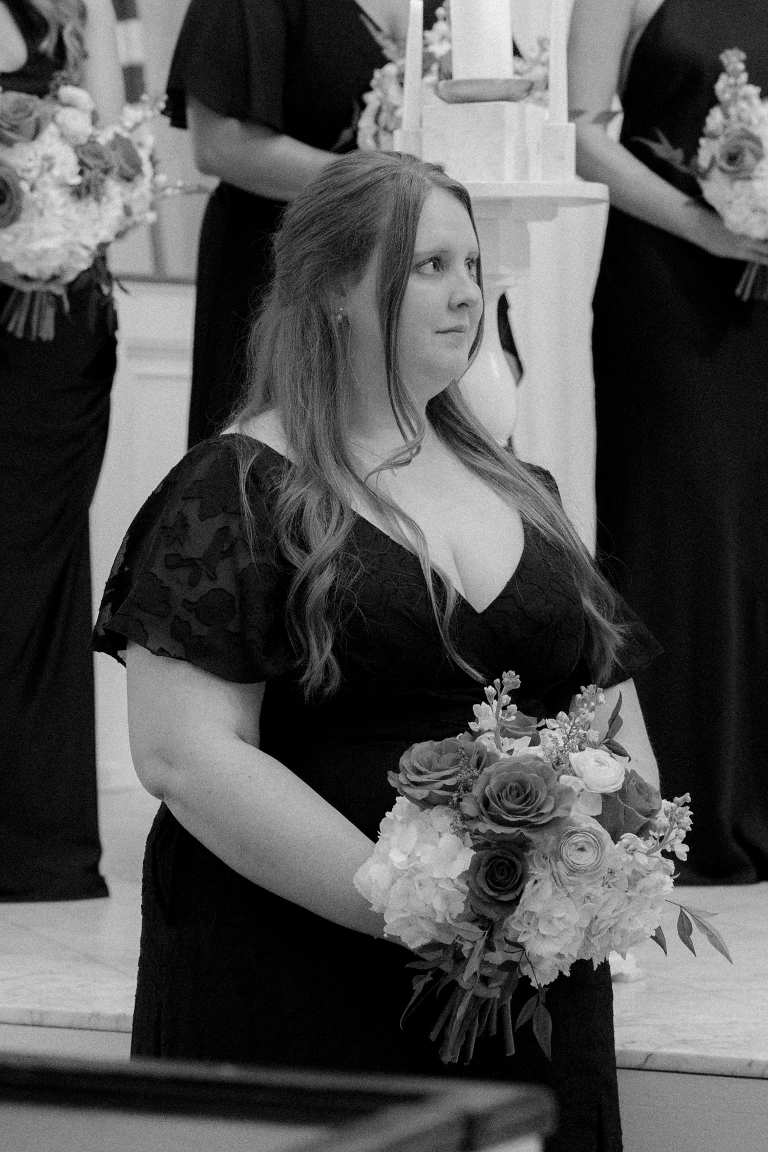 Bridesmaid in black lace dress holding floral bouquet during wedding ceremony, black and white photo.