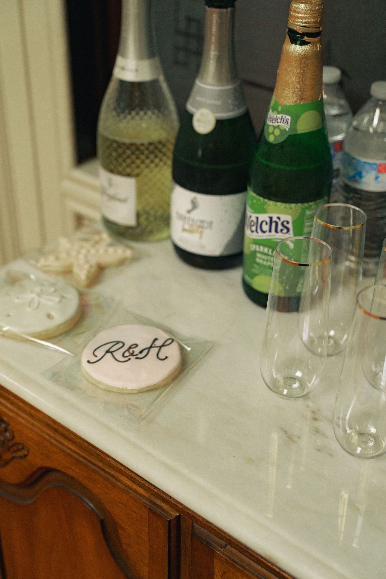 Wedding cookie with R&H monogram beside champagne bottles and flutes on marble sideboard.
