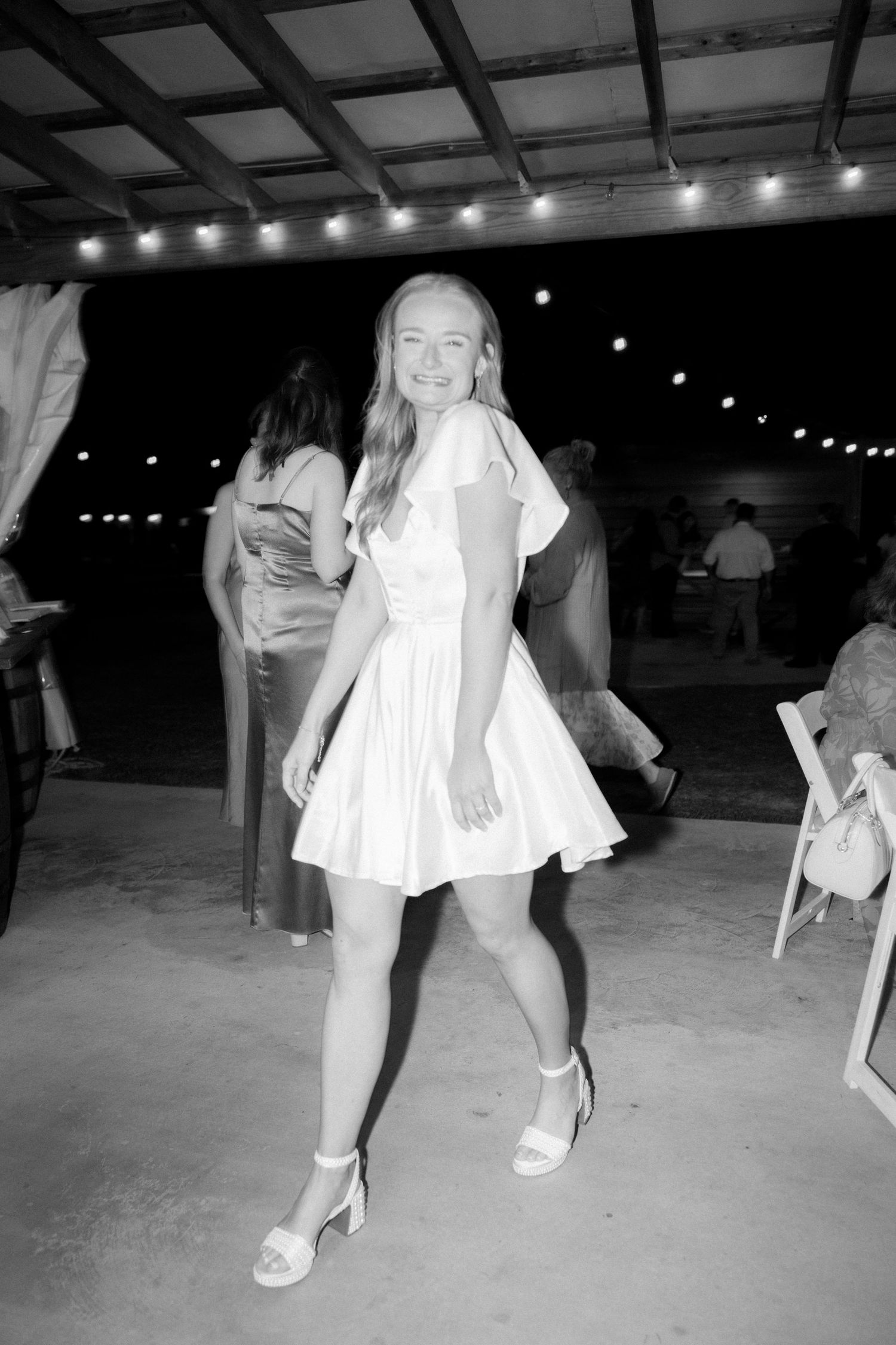 Black and white photo of smiling woman in white mini dress dancing at outdoor wedding reception.