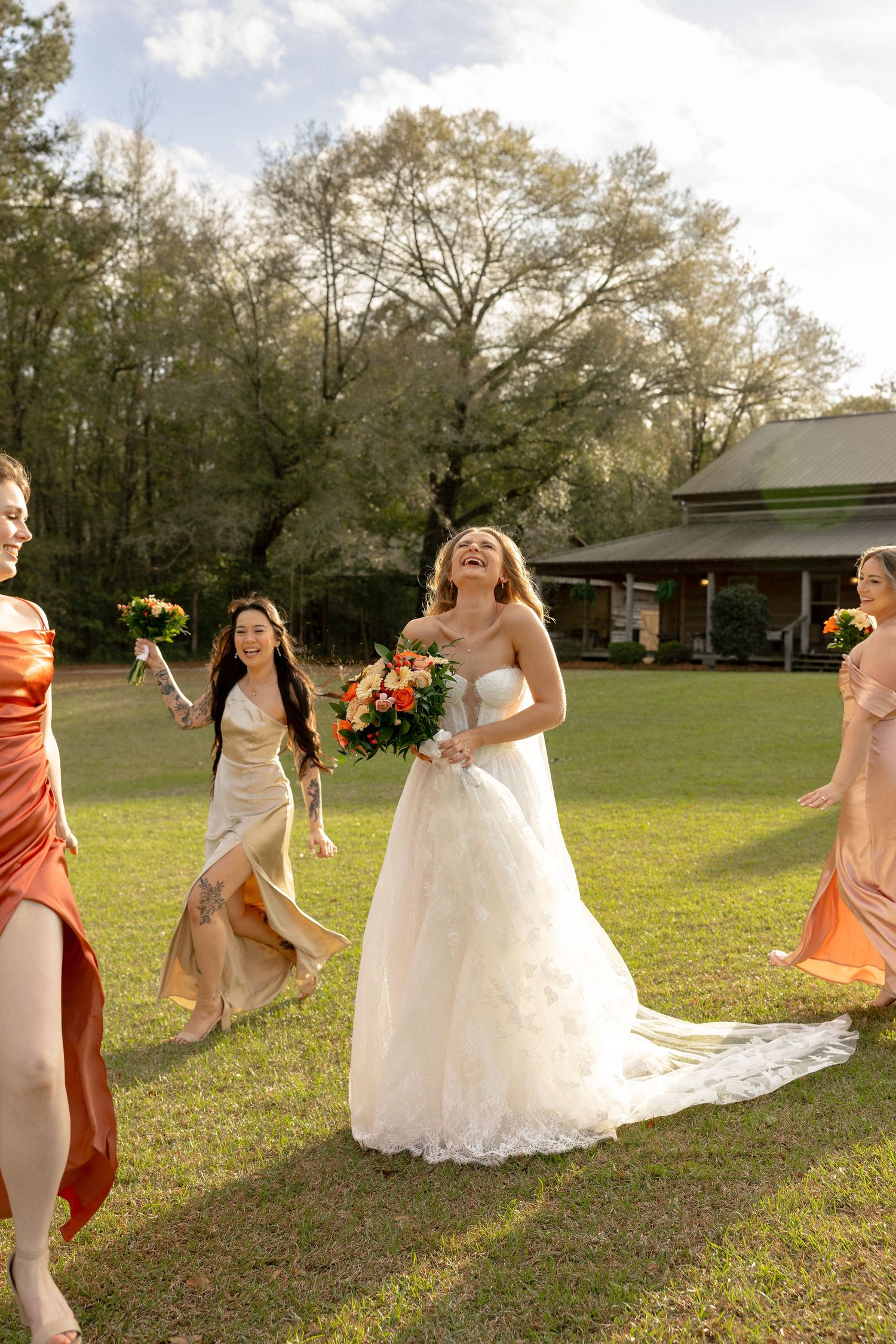 Laughing bride in white ballgown holding orange bouquet walking with bridesmaids on sunny green lawn.