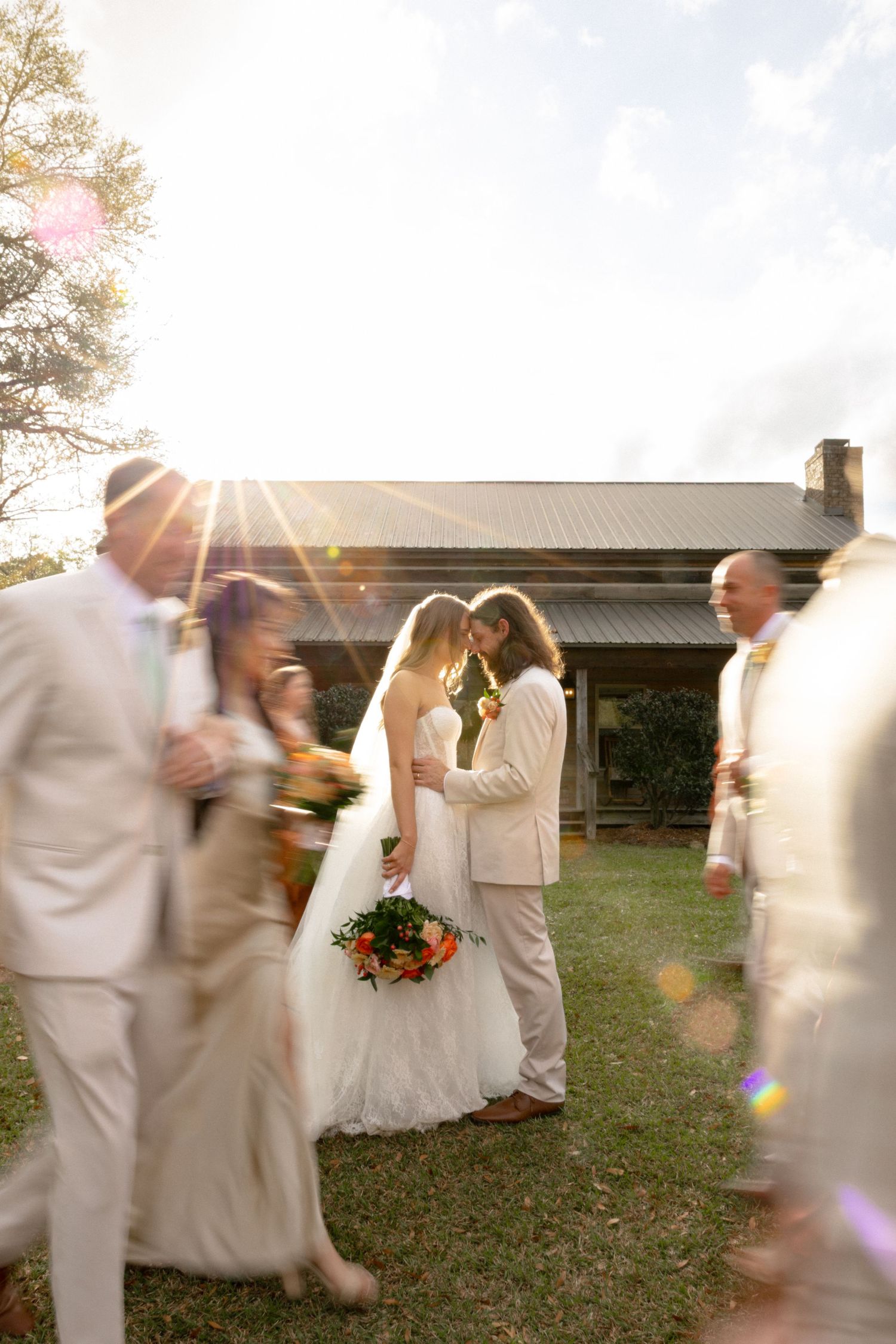 Bride and groom share intimate moment surrounded by moving wedding guests in sunlight.