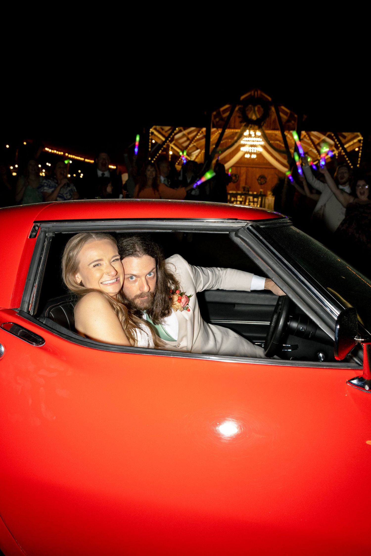 A couple smiles in a red classic car at night with colorful festival lights glowing in the background.