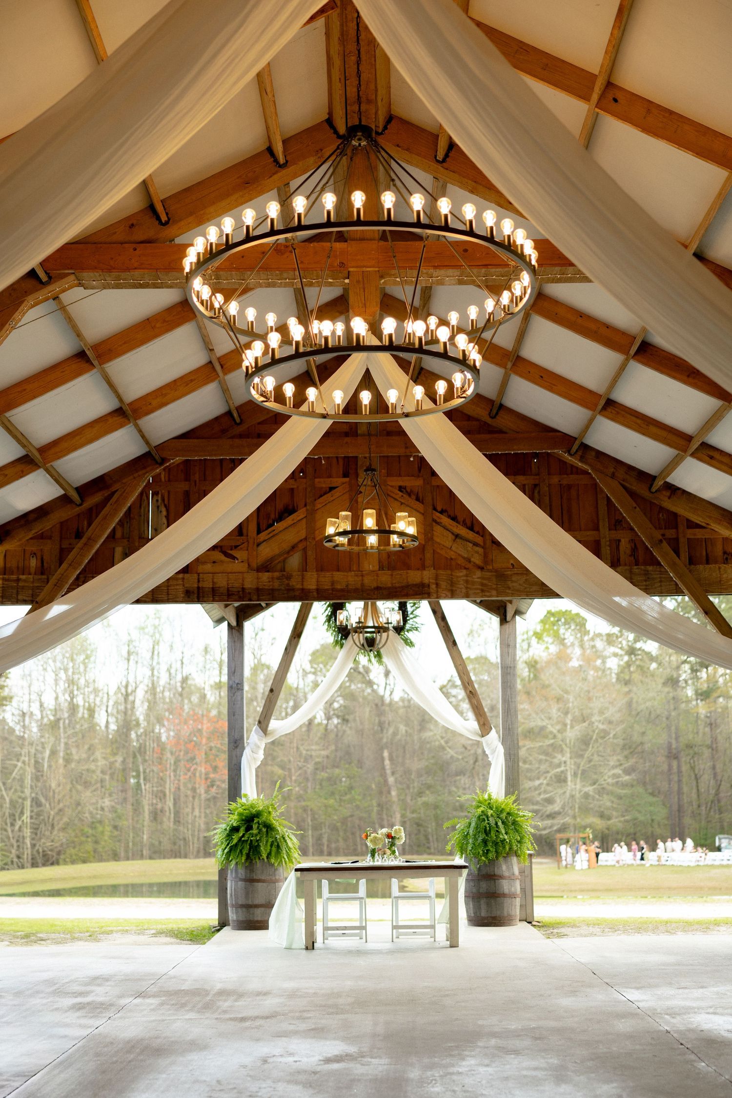 Rustic barn wedding venue with grand chandelier, draped fabric, and sweetheart table setup.