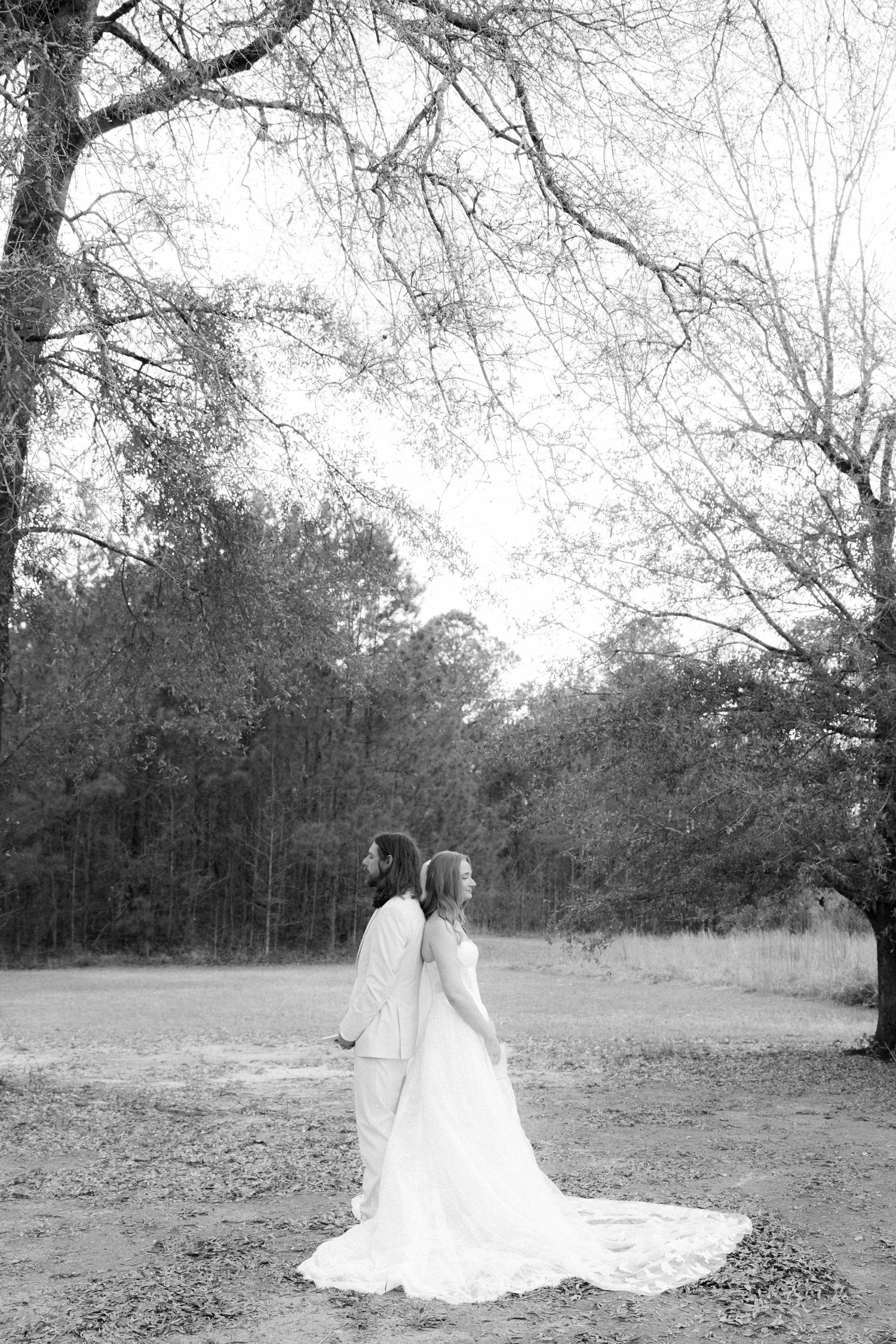 Bride and groom stand back to back in wooded outdoor setting with long white dress train in black and white.