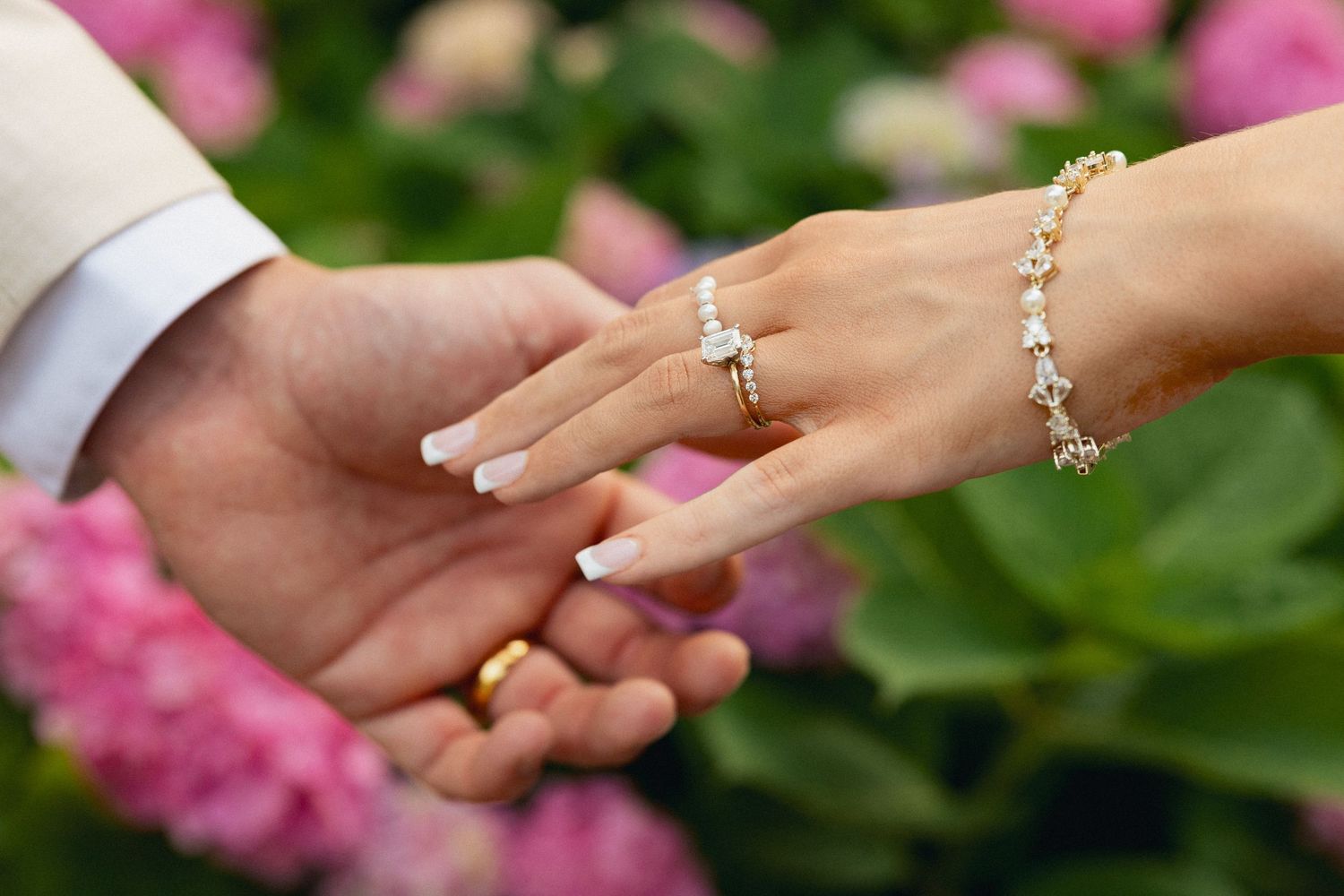 Close-up of bride and groom holding hands showcasing diamond ring and bracelet with pink flowers.