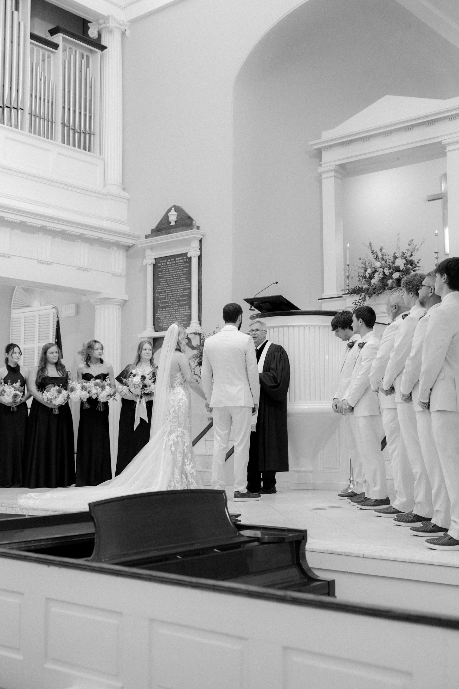 Bride and groom hold hands at altar flanked by bridesmaids in black and groomsmen in white suits.
