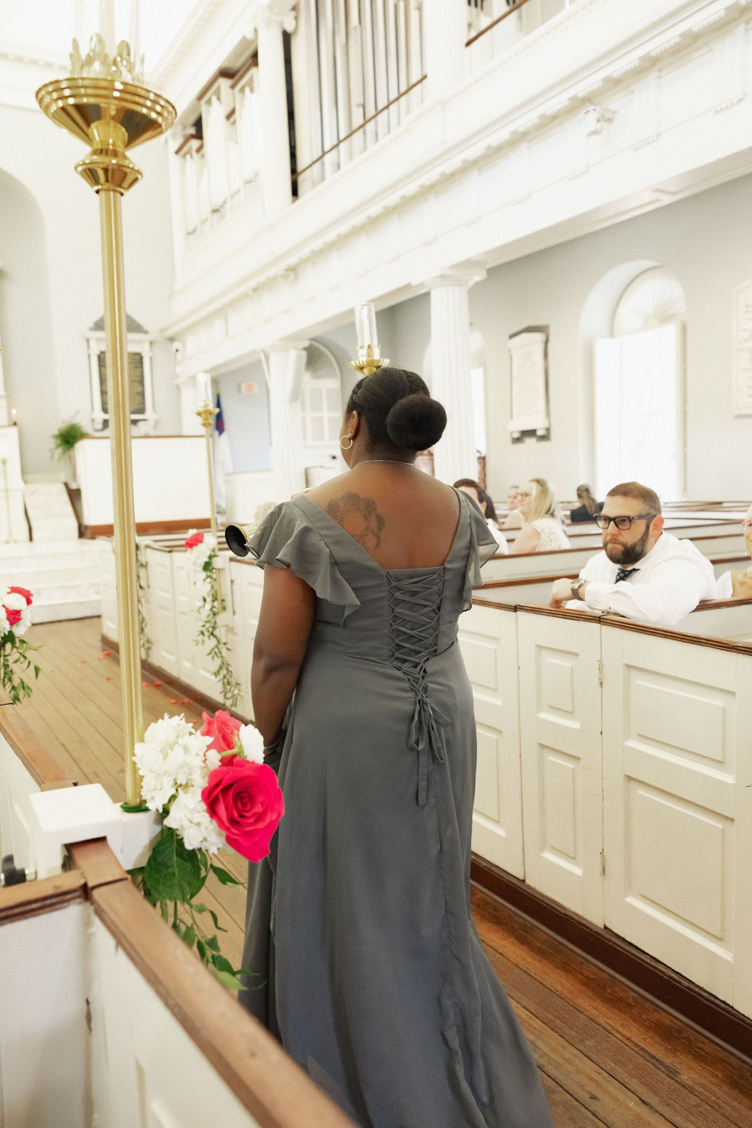 Bridesmaid in gray gown holds pink and white roses while escorting guests to pews in a classic church.