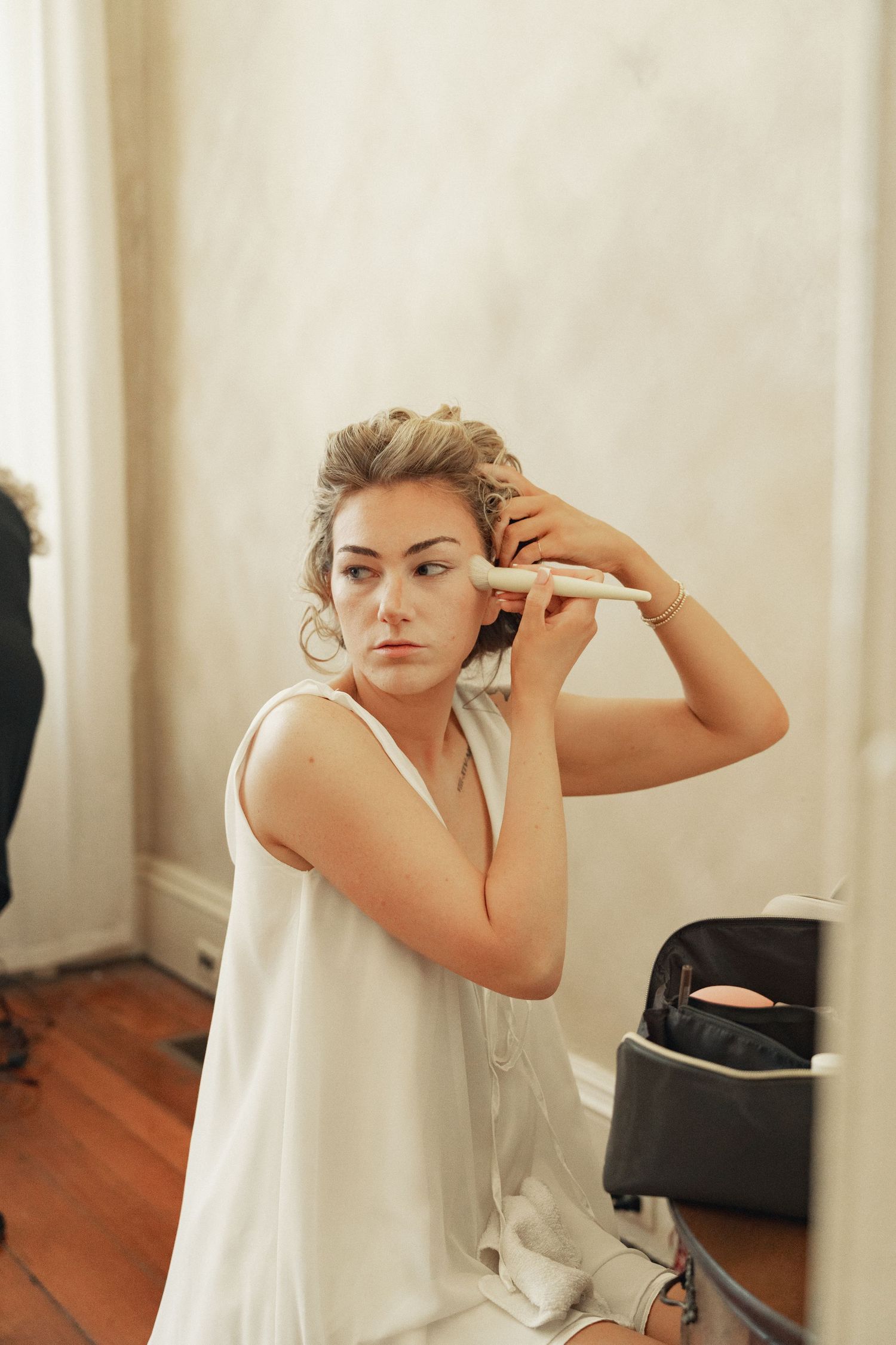 A bride in a white dress curls her updo hairstyle with a curling iron while preparing for her wedding.