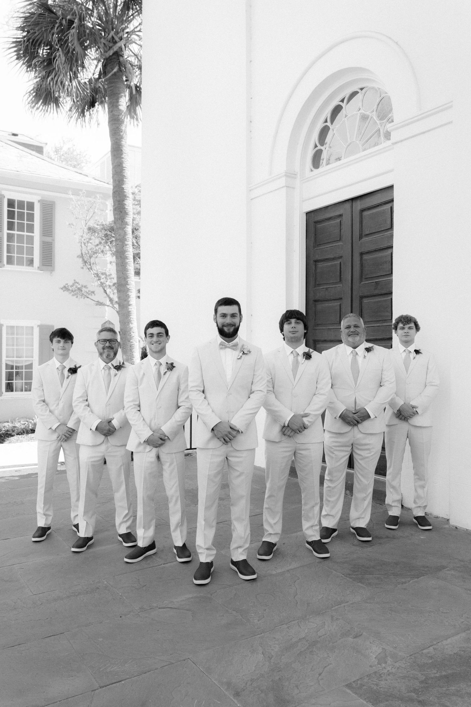 Groom and groomsmen pose in matching white suits outside a white church in black and white wedding photo.