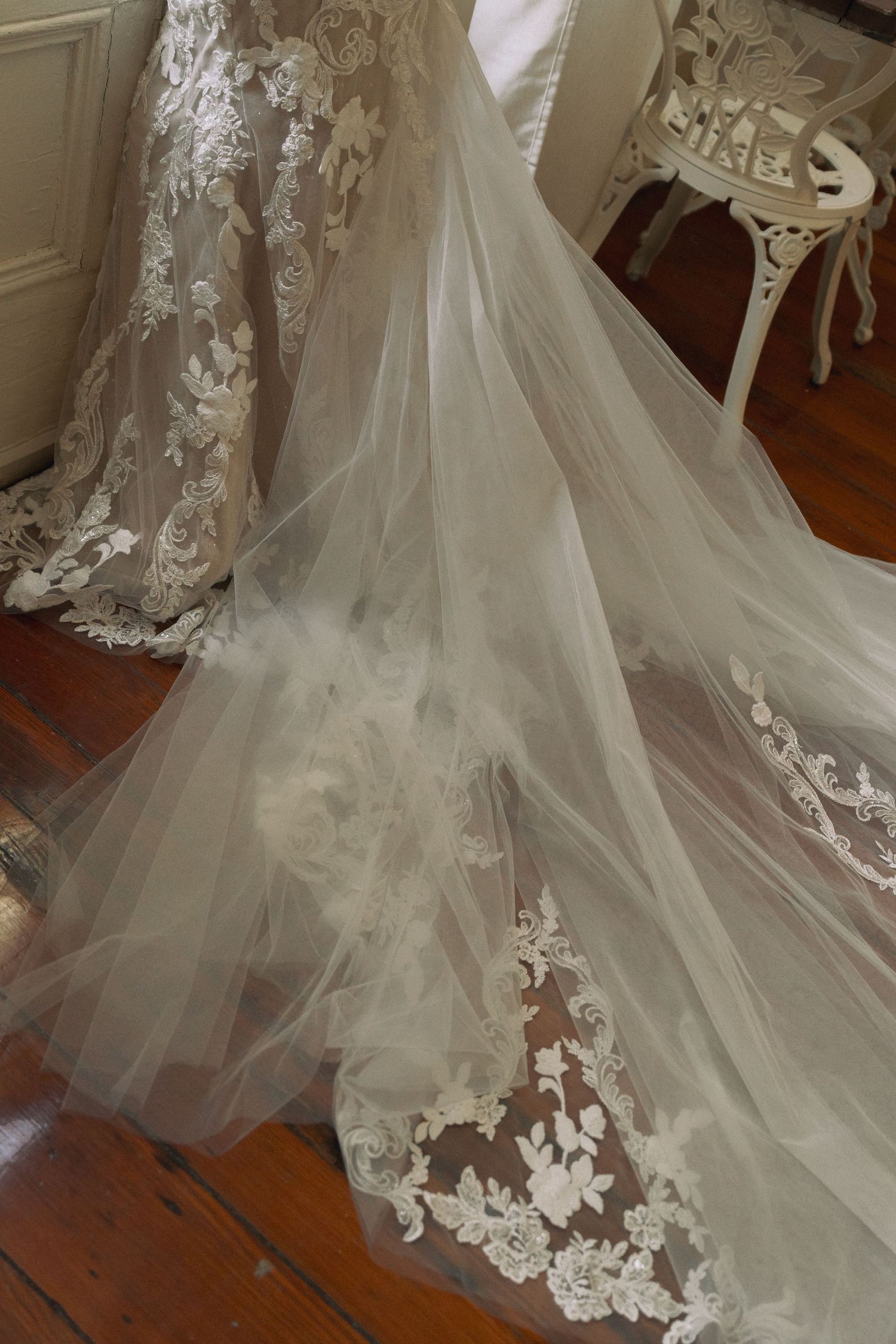 Elegant wedding dress with intricate lace appliqués and flowing tulle train displayed on hardwood floor.