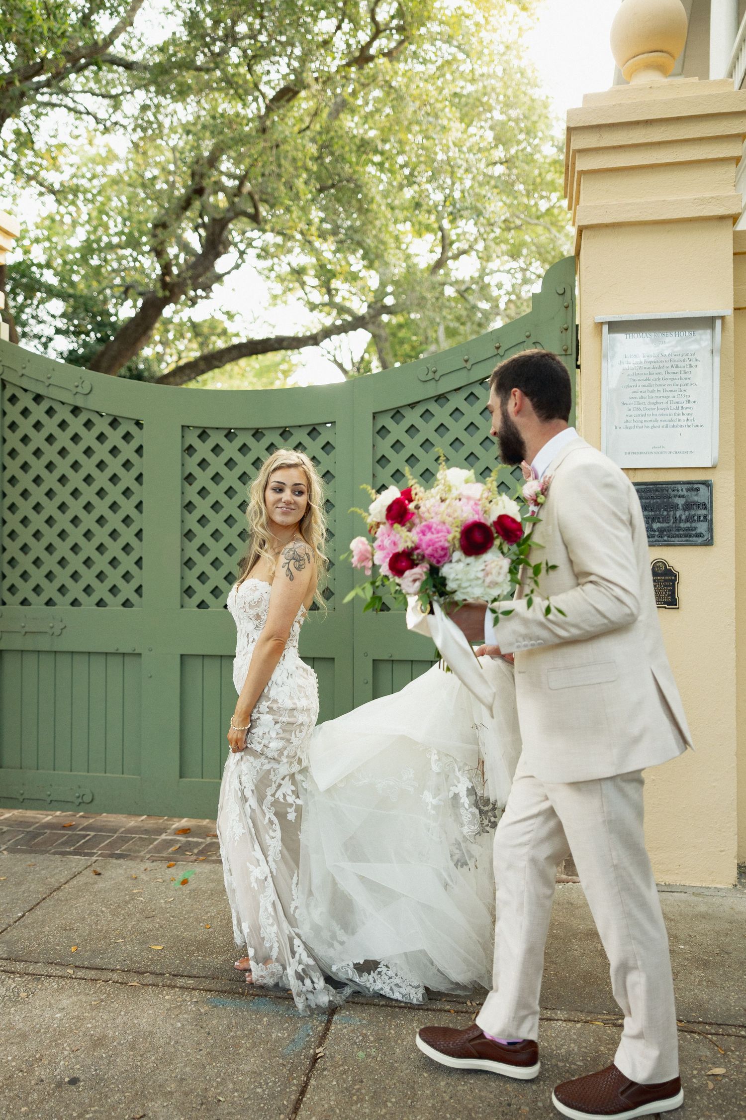 Groom presenting a vibrant pink and red bridal bouquet to his smiling bride at a green garden gate.