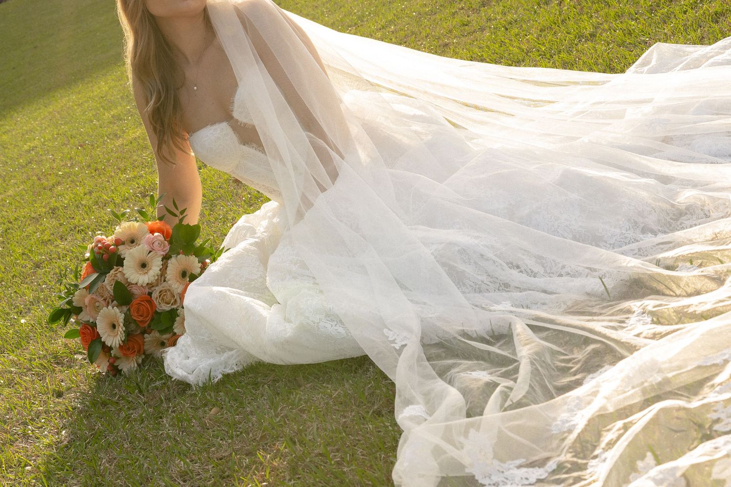 Bride in flowing lace wedding gown with long veil bending to pick up colorful bouquet on grass.