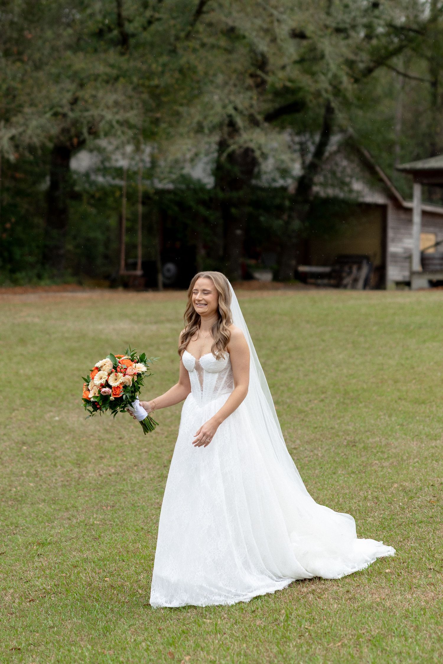 Radiant bride in white strapless gown with cathedral veil holding orange bouquet on green lawn.
