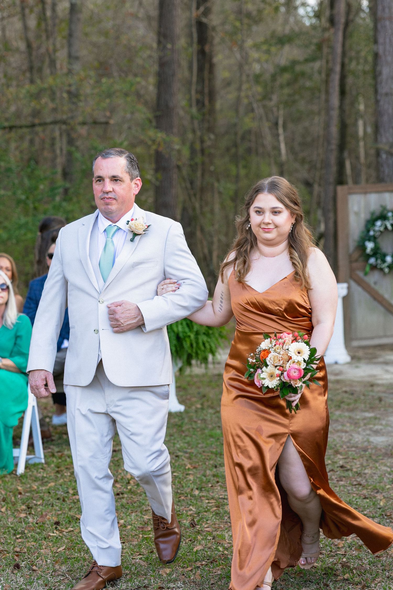 A groomsman in gray suit with mint tie escorts a smiling bridesmaid in copper satin gown down an outdoor wedding aisle.