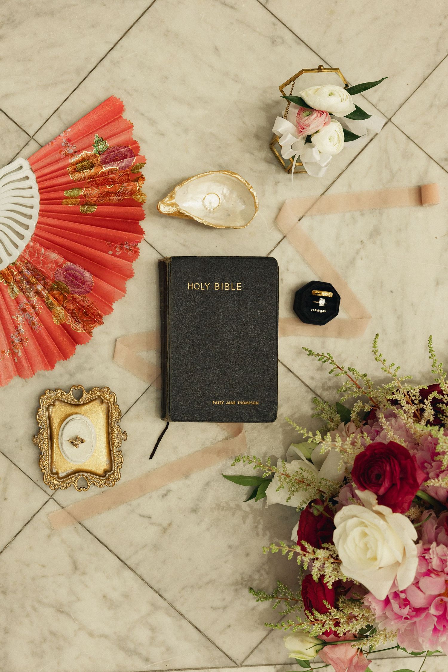 Wedding flat lay with Holy Bible, floral bouquet, red fan, ribbon, ring box, and gold frame on marble surface.