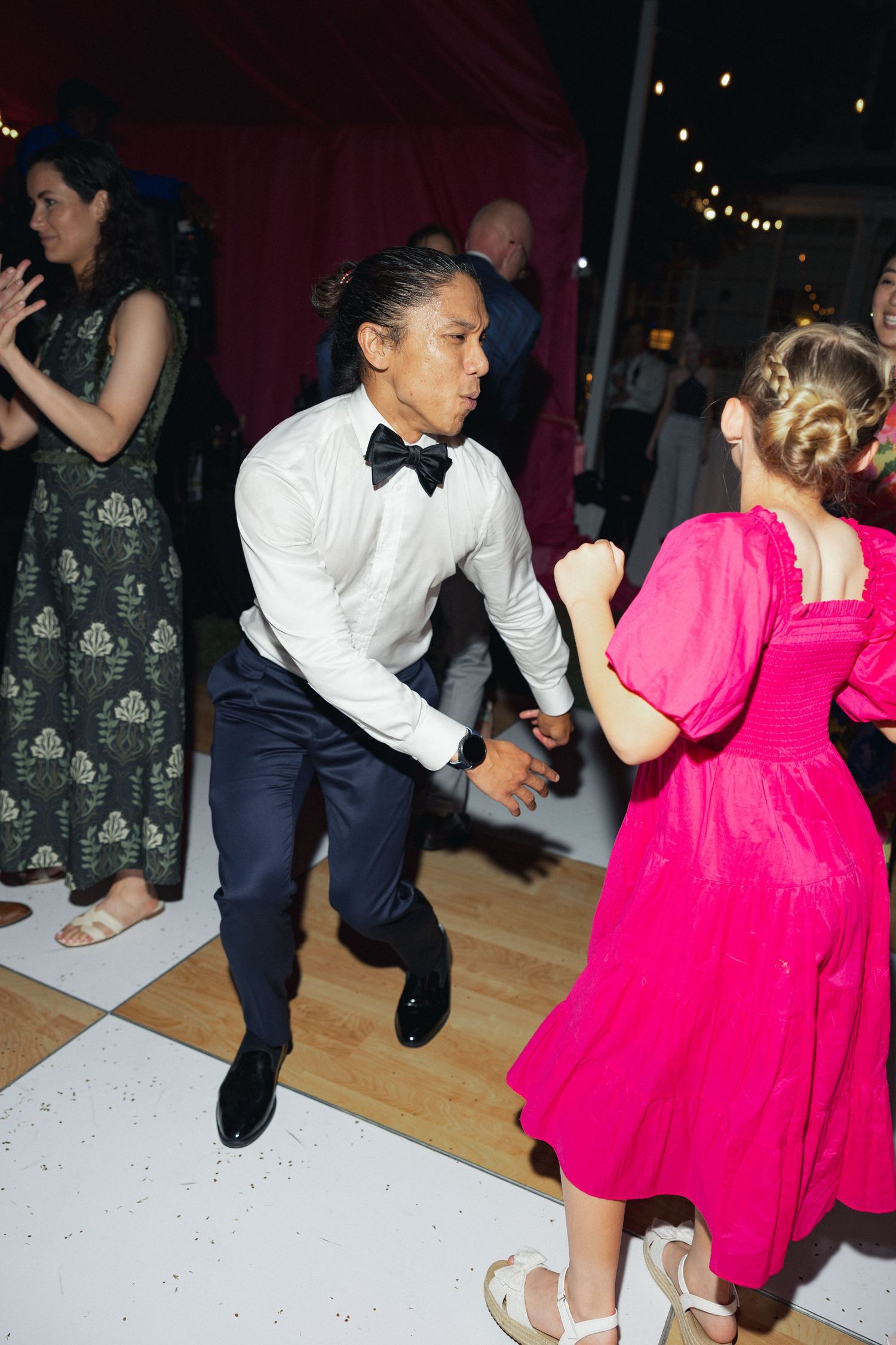 A man in a white shirt and bow tie dances energetically with a woman in a pink dress at a wedding reception.