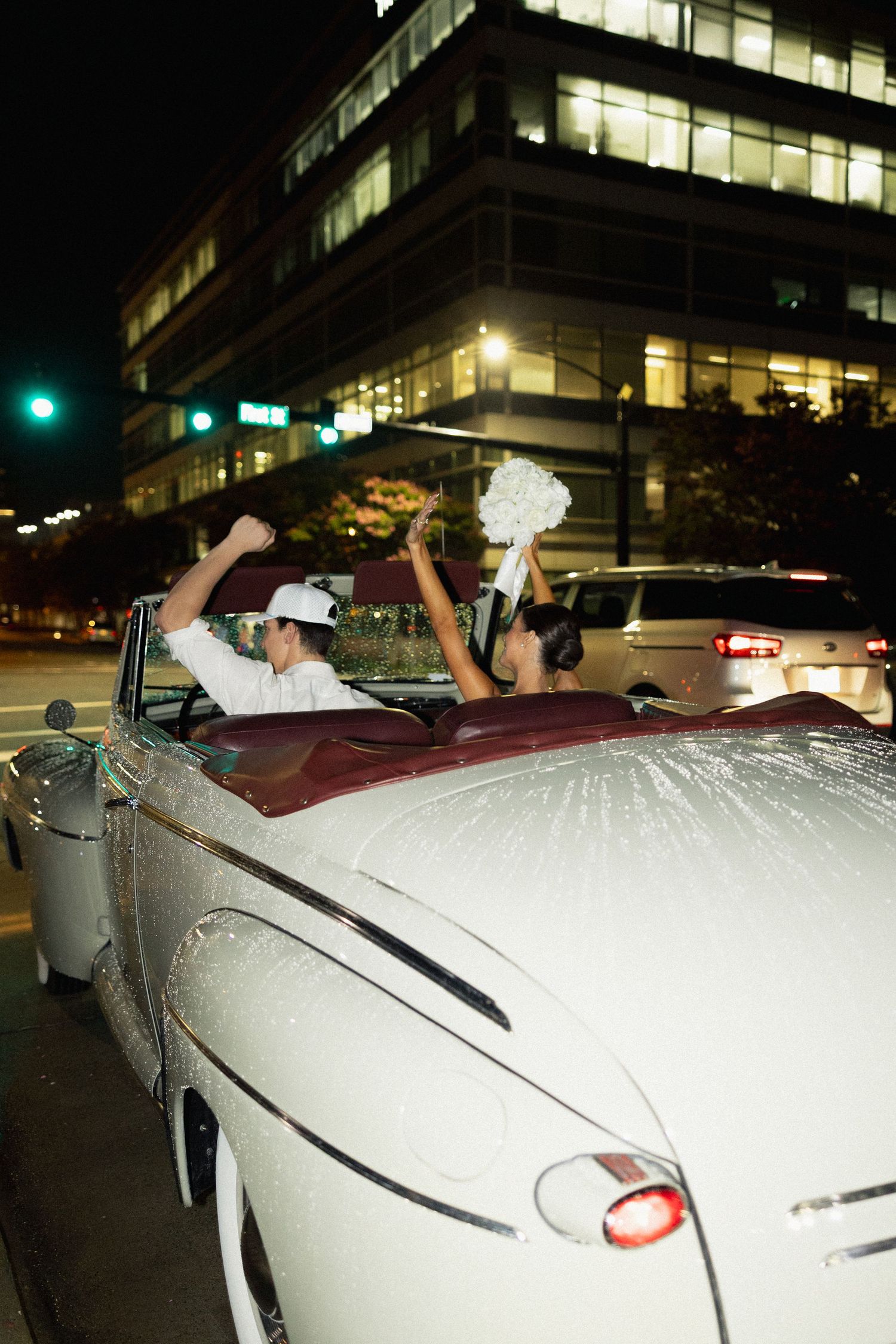 Newlyweds celebrating in a classic white convertible car at night, bride holding bouquet aloft.