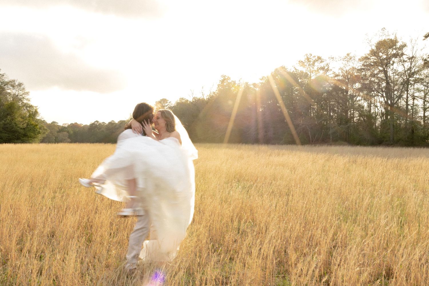 Groom lifting bride in a romantic kiss in a golden sunlit field at sunset.