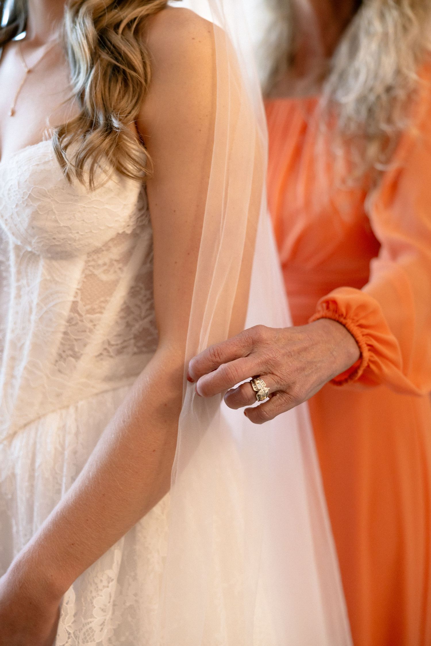 Close-up of mother in orange dress holding bride's hand in lace wedding gown with veil.