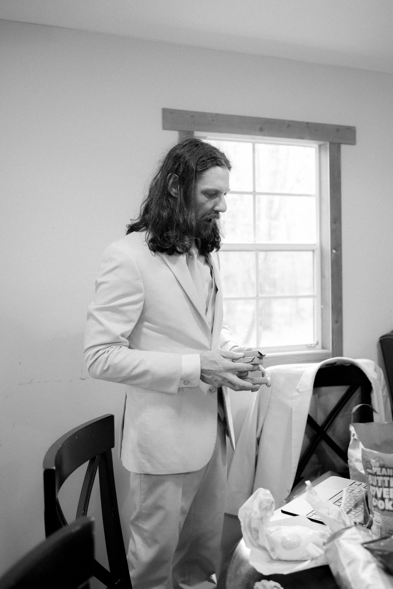 Black and white photo of long-haired bearded groom in light suit preparing before wedding ceremony.