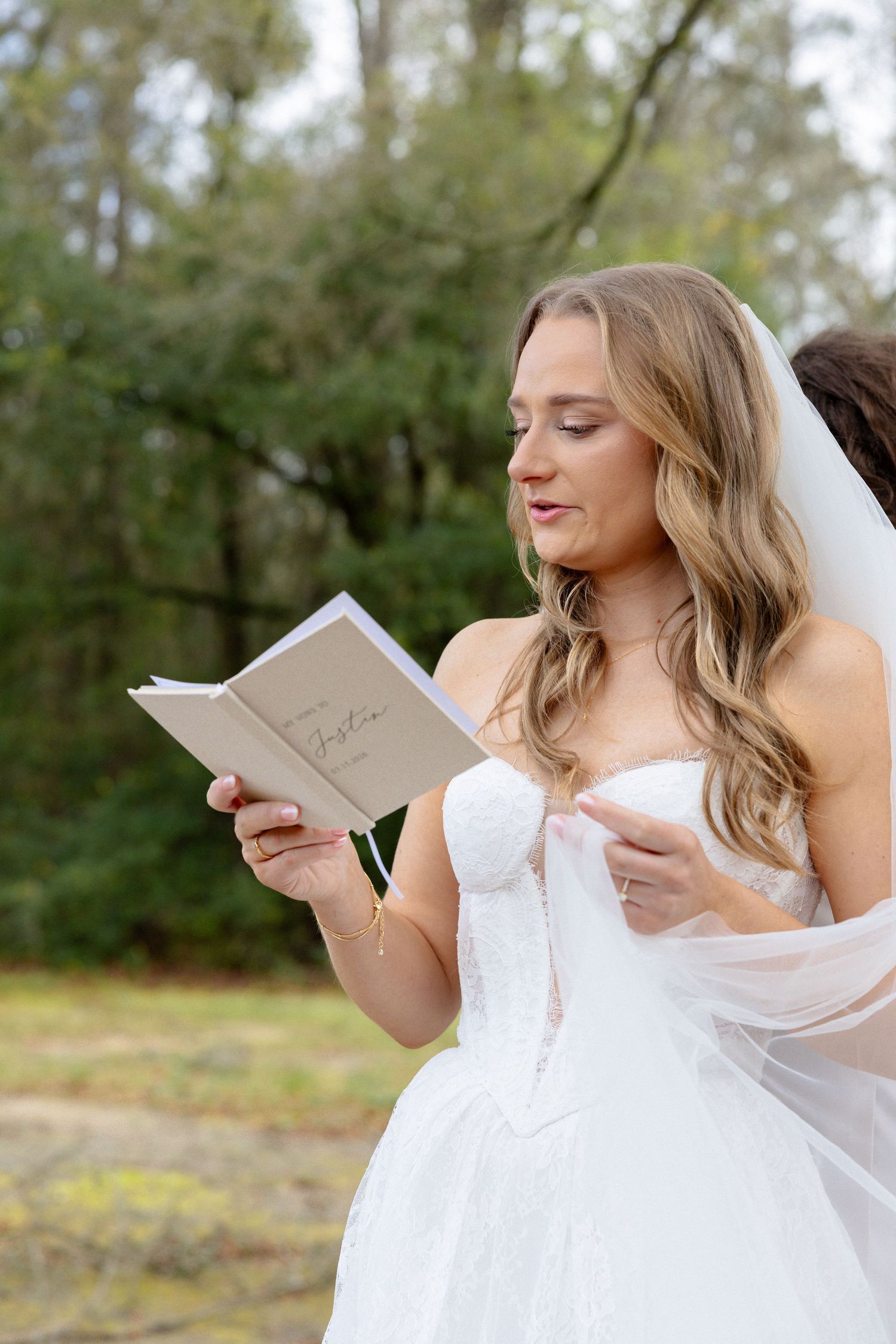 Blonde bride in strapless lace wedding gown reads from small book with veil flowing in outdoor setting.
