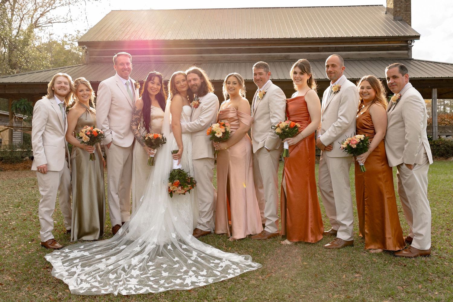 Large wedding party poses together in warm sunlight outside rustic wooden venue.
