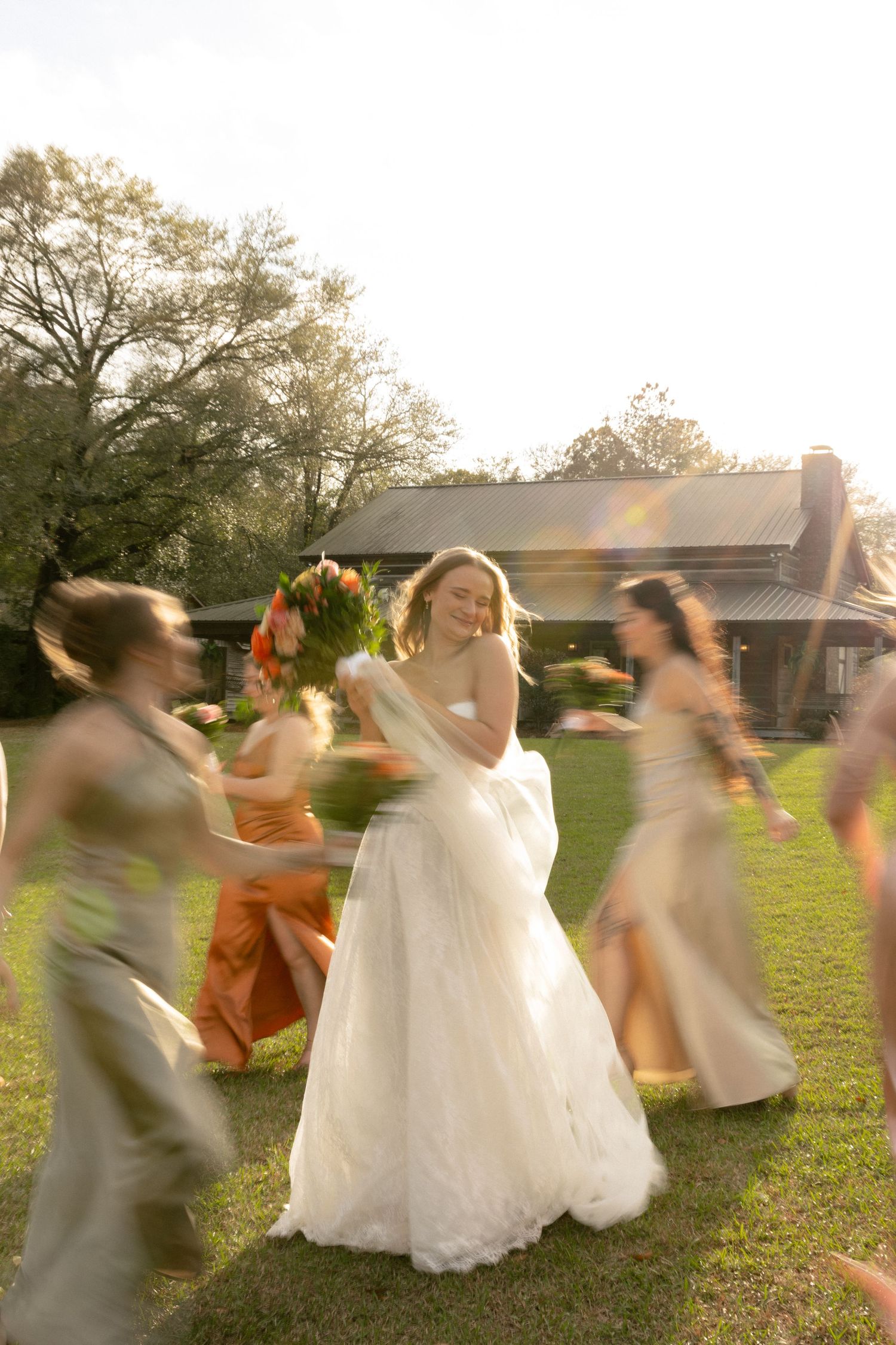 Motion blur photo of bride in white dress dancing with bridesmaids in sage and orange gowns outdoors.