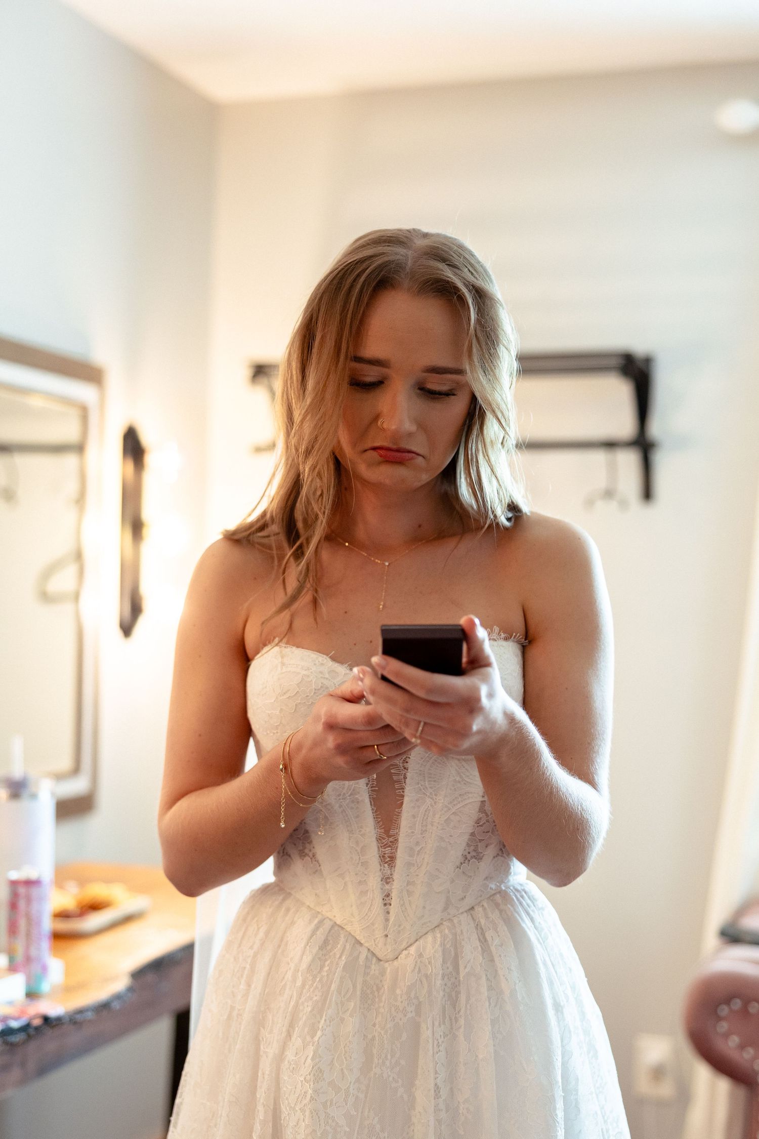 Bride in white lace wedding dress looking at her phone with a concerned expression before the ceremony.