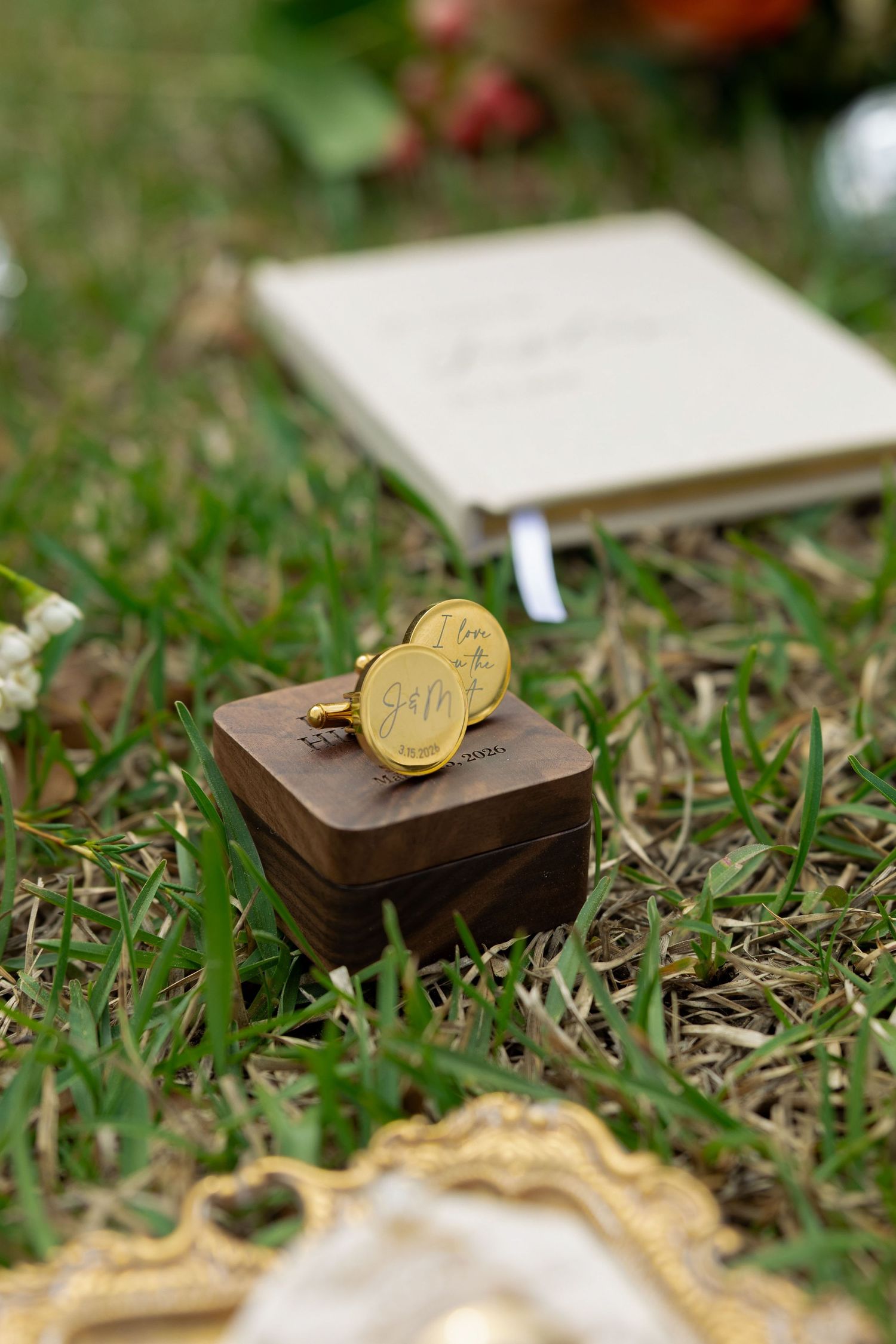 Gold engraved wedding ring displayed on a wooden ring box in green grass with a journal in the background.