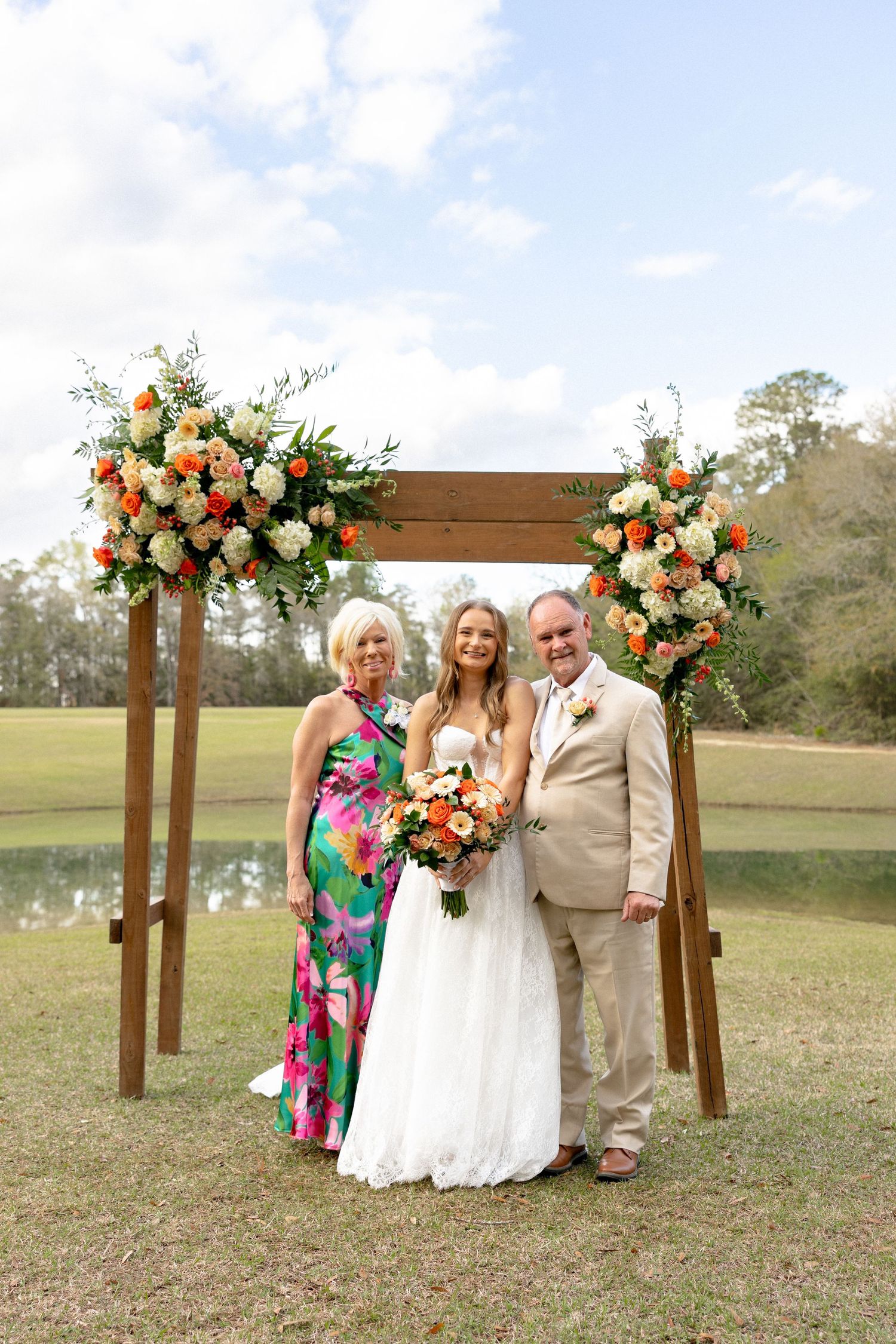 Bride with parents under wooden arch adorned with orange and white flowers outdoors.