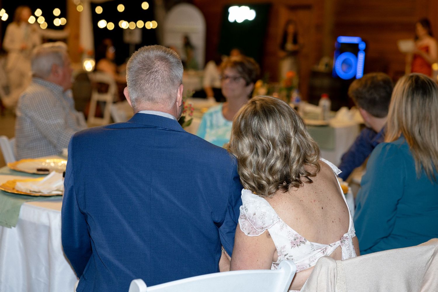 Bride in white lace gown and groom in blue suit sit together at wedding reception dinner table.