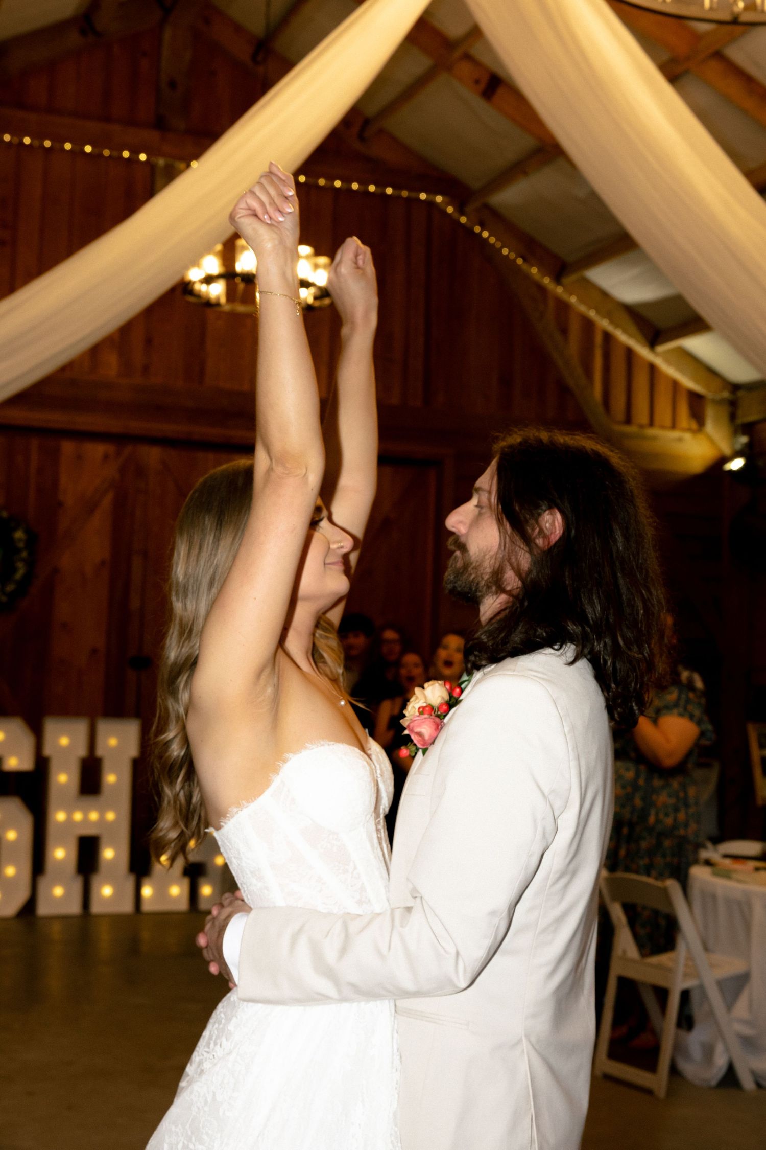 Bride and groom share their first dance as groom twirls bride under draped ceiling lights in barn venue.