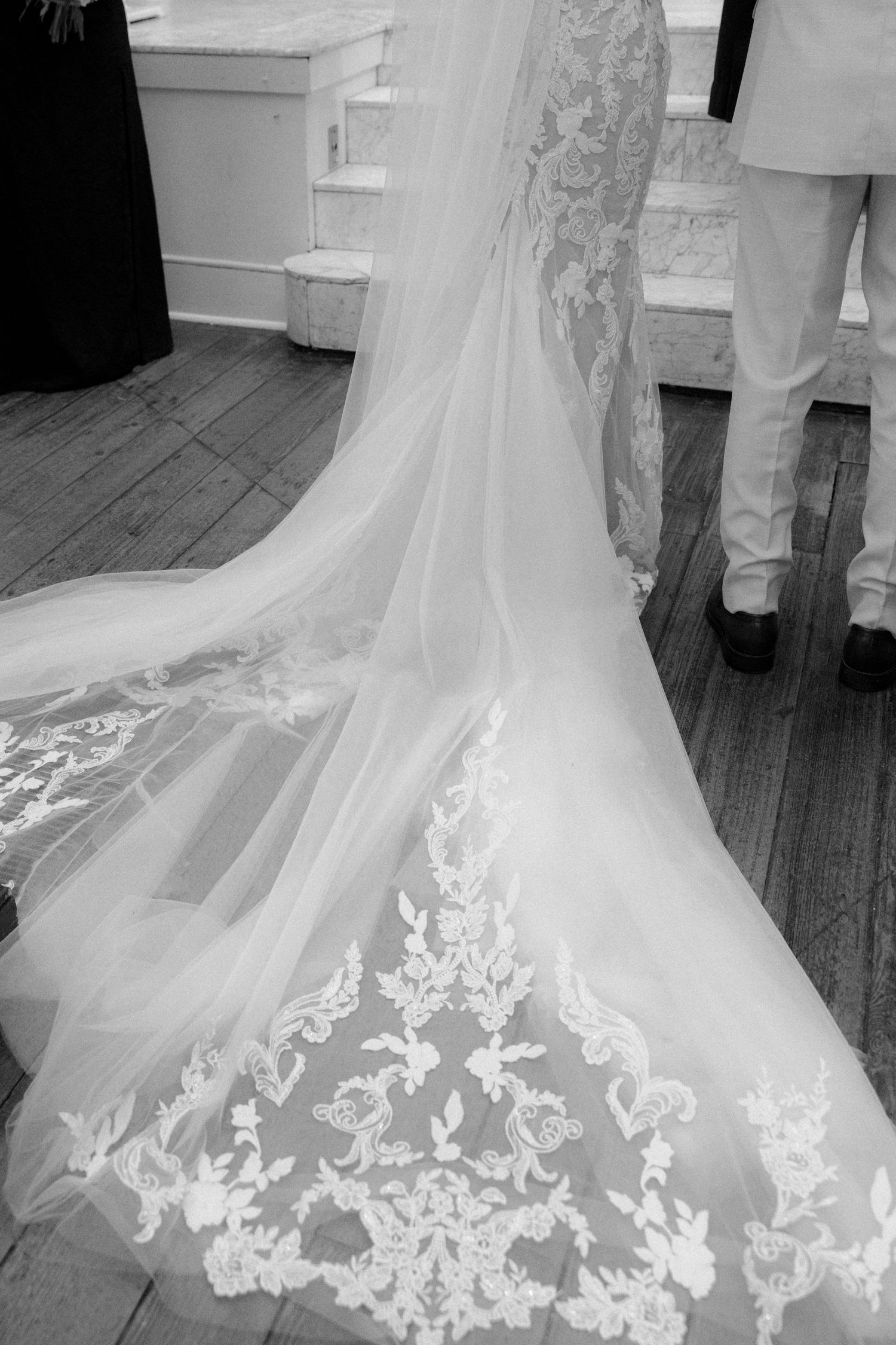 Close-up of an intricate lace wedding dress train spread across a wooden church floor.