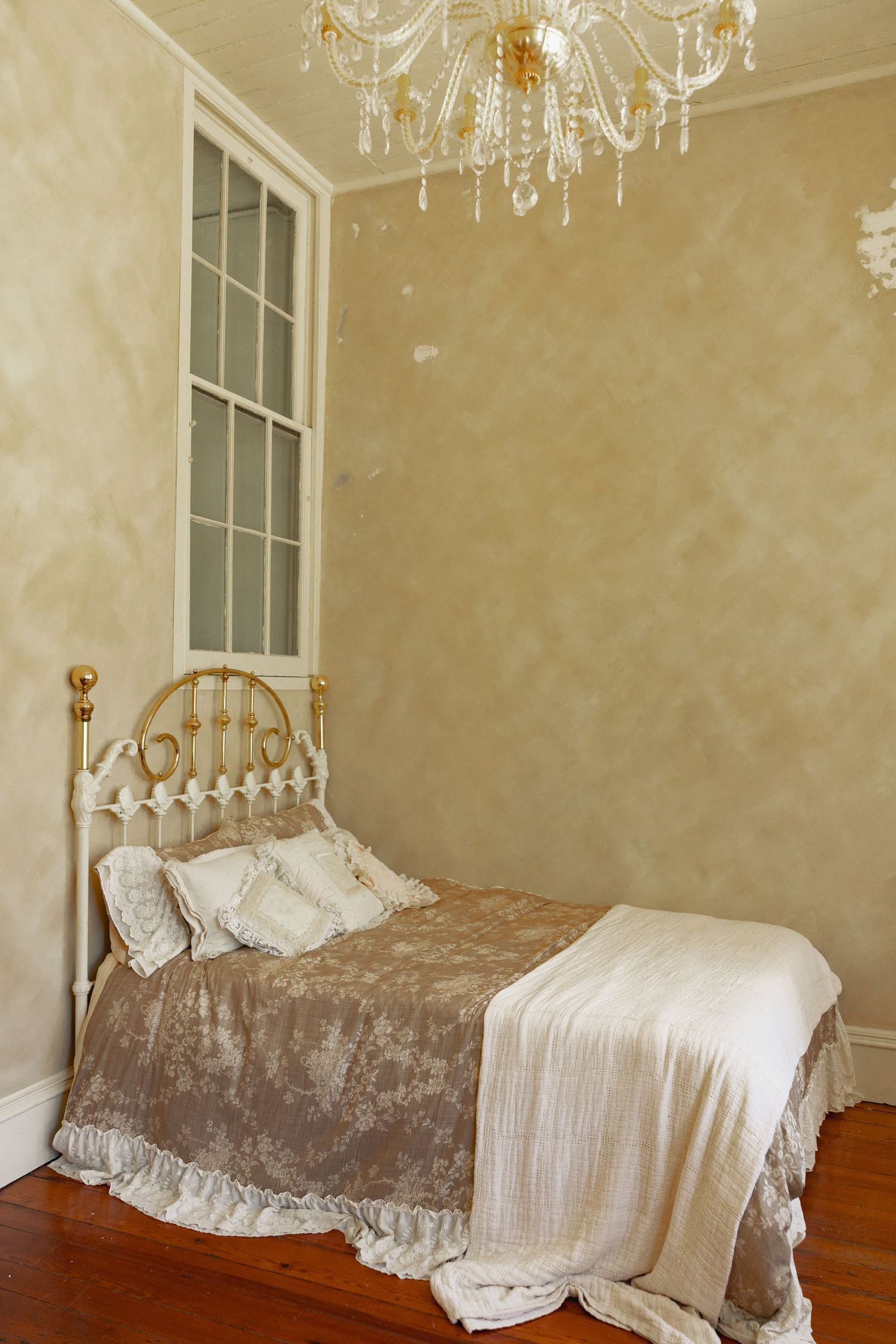 Elegant vintage bedroom with brass bed, crystal chandelier, damask bedding, and textured beige walls on hardwood floors.