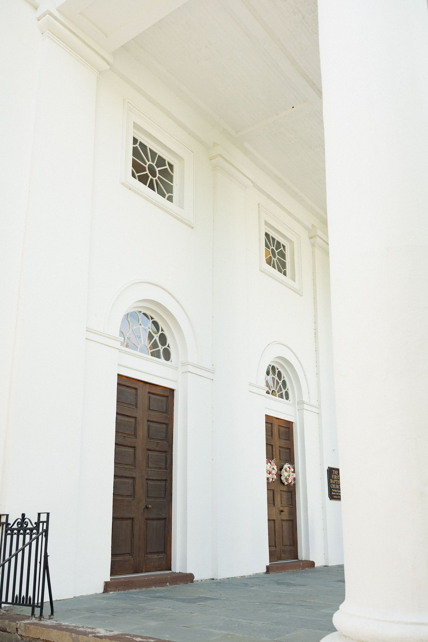 White colonial church facade with two brown wooden doors, arched windows, and ornate stained glass details.