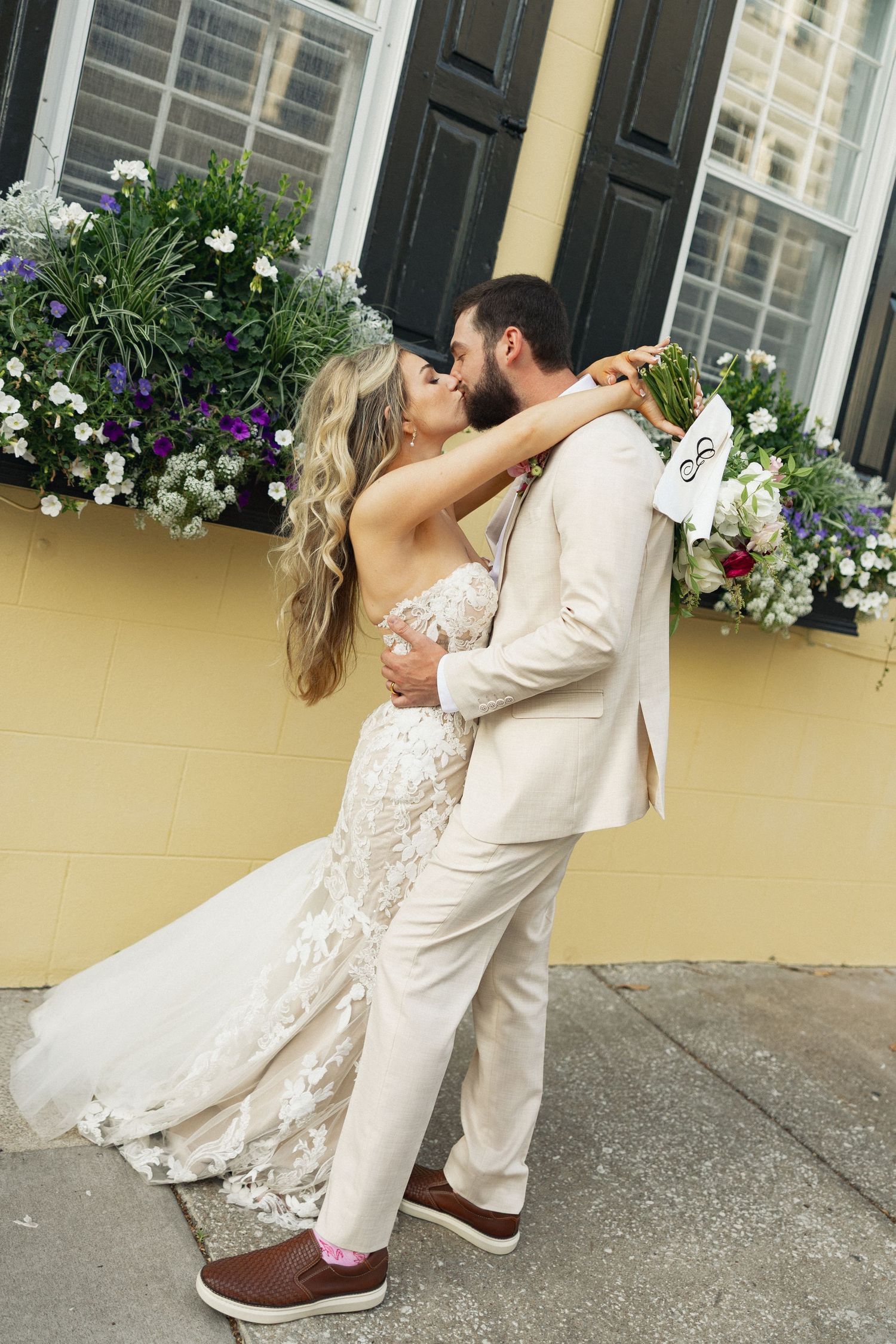 Bride and groom share a passionate kiss in front of a yellow building adorned with colorful flower boxes.