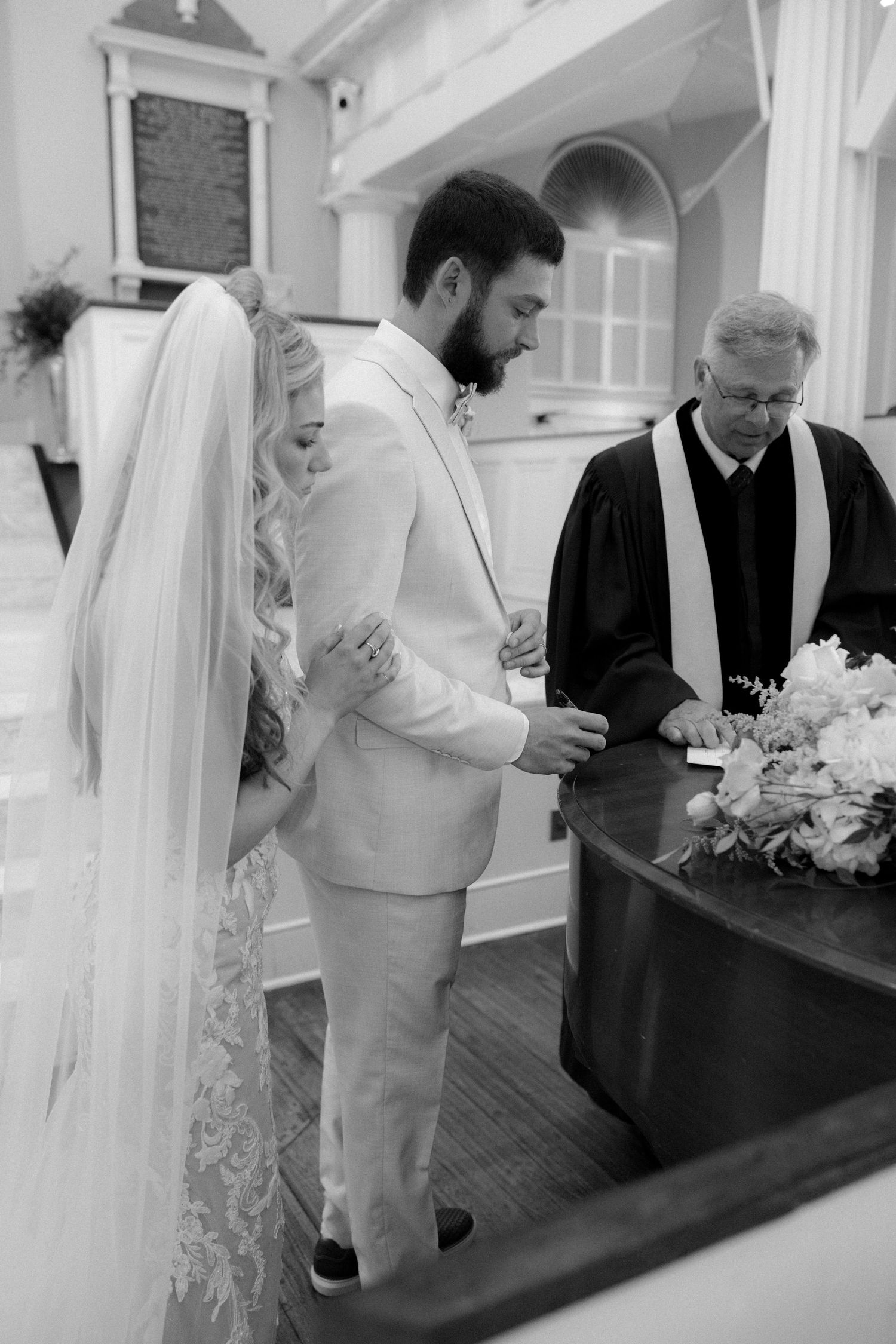 Black and white photo of a bride and groom bowing in prayer with an officiant during a church wedding ceremony.