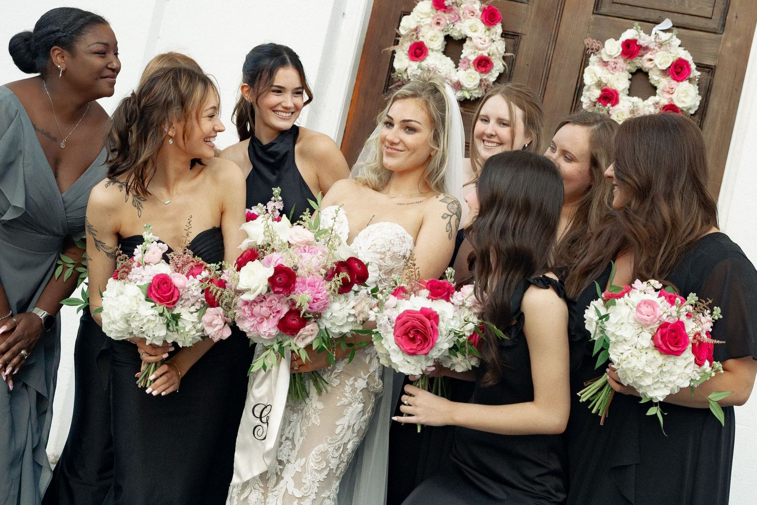 Bride surrounded by bridesmaids in black dresses holding pink and white floral bouquets.