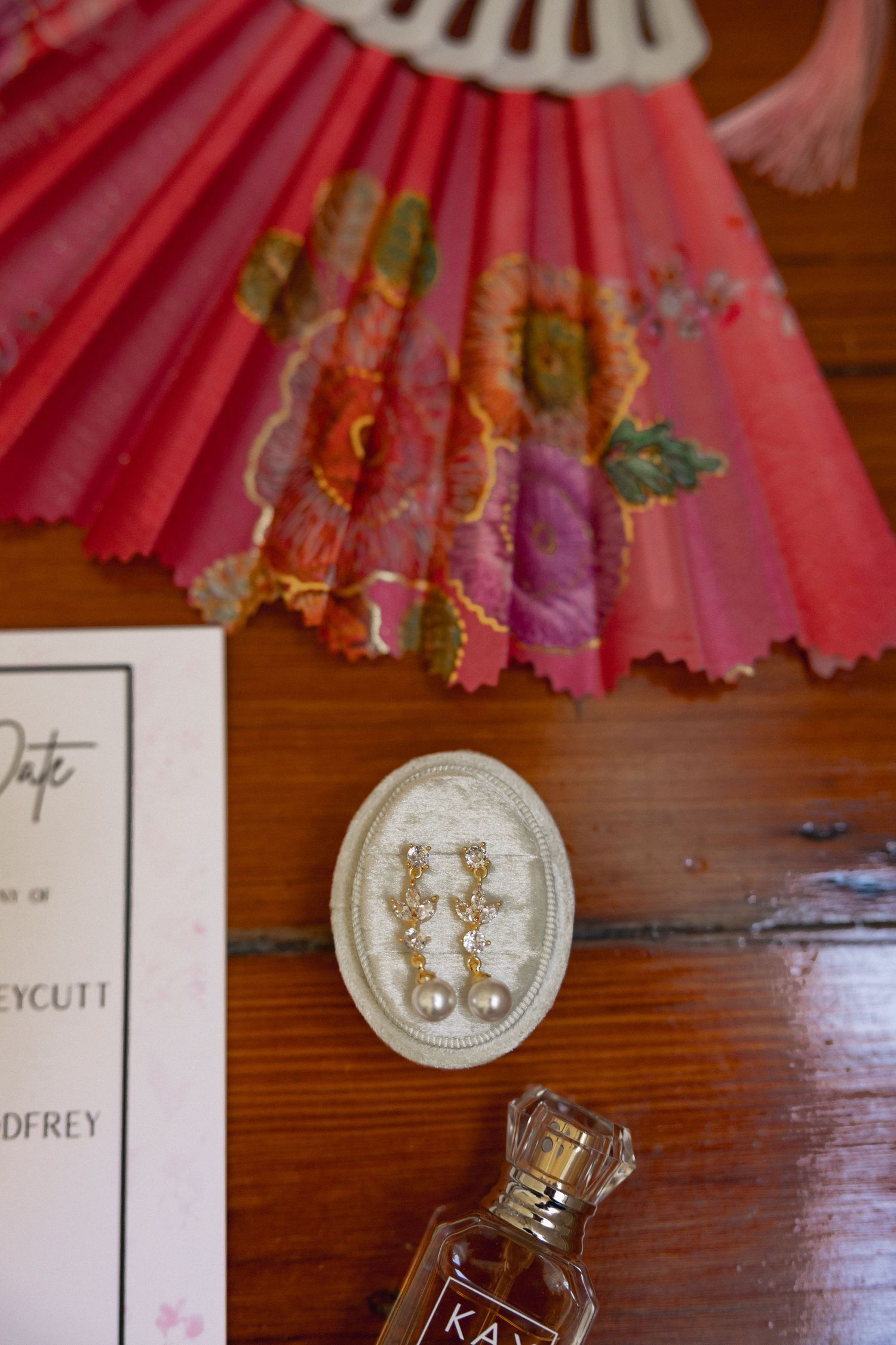 Pink floral folding fan with pearl drop earrings on oval tray, perfume bottle, and wedding save the date card on wood table.
