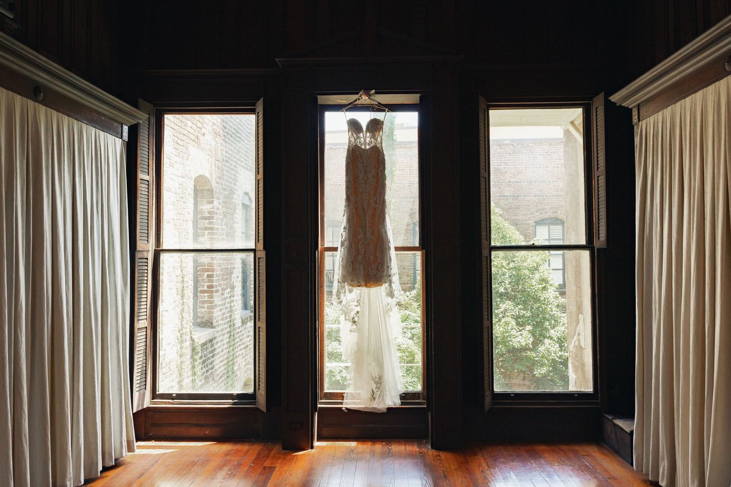 Wedding dress hanging in a sunlit window of a dark wood-paneled room with hardwood floors.