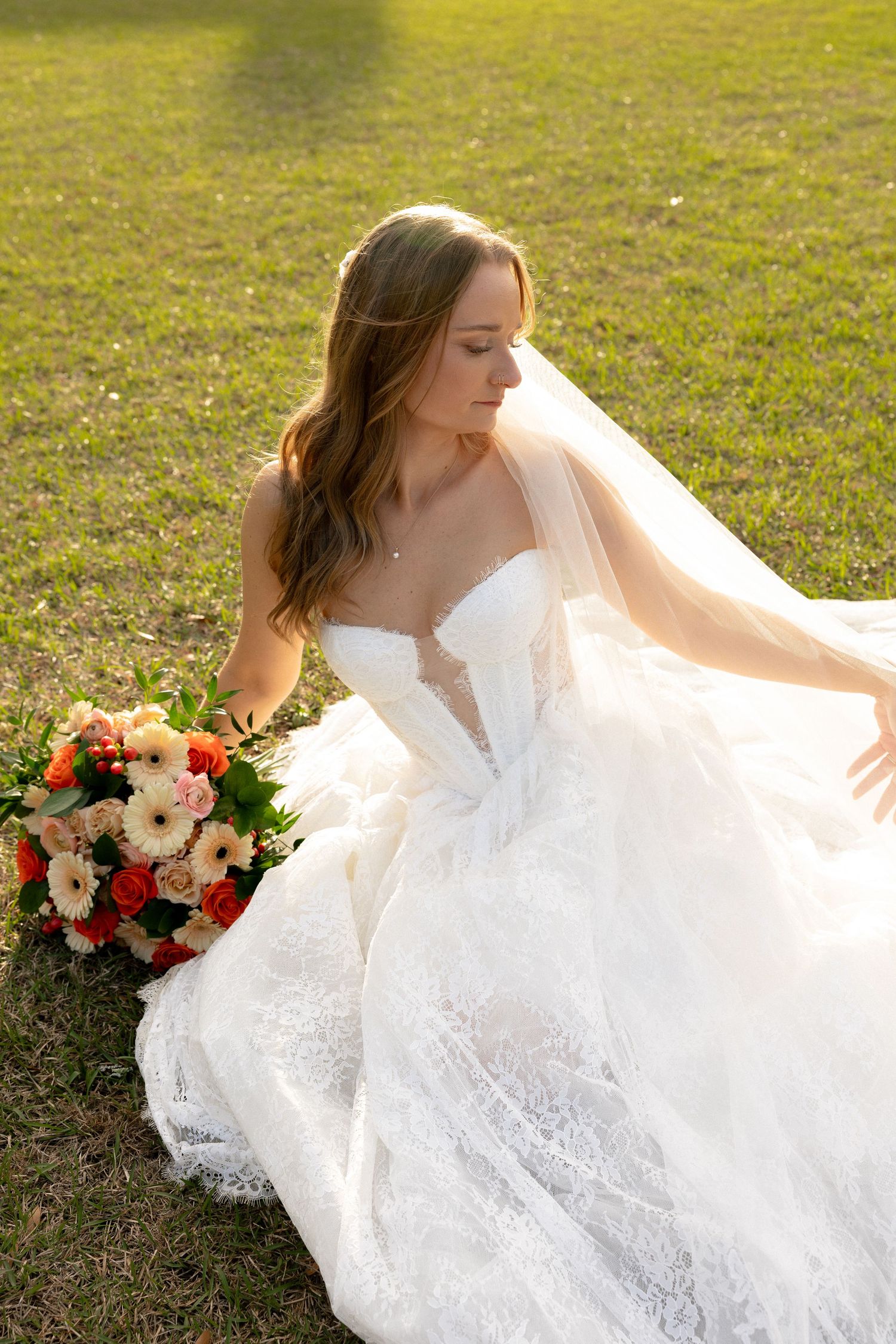 Bride in white lace ballgown sits on green grass holding colorful bouquet of orange and pink flowers in golden sunlight.