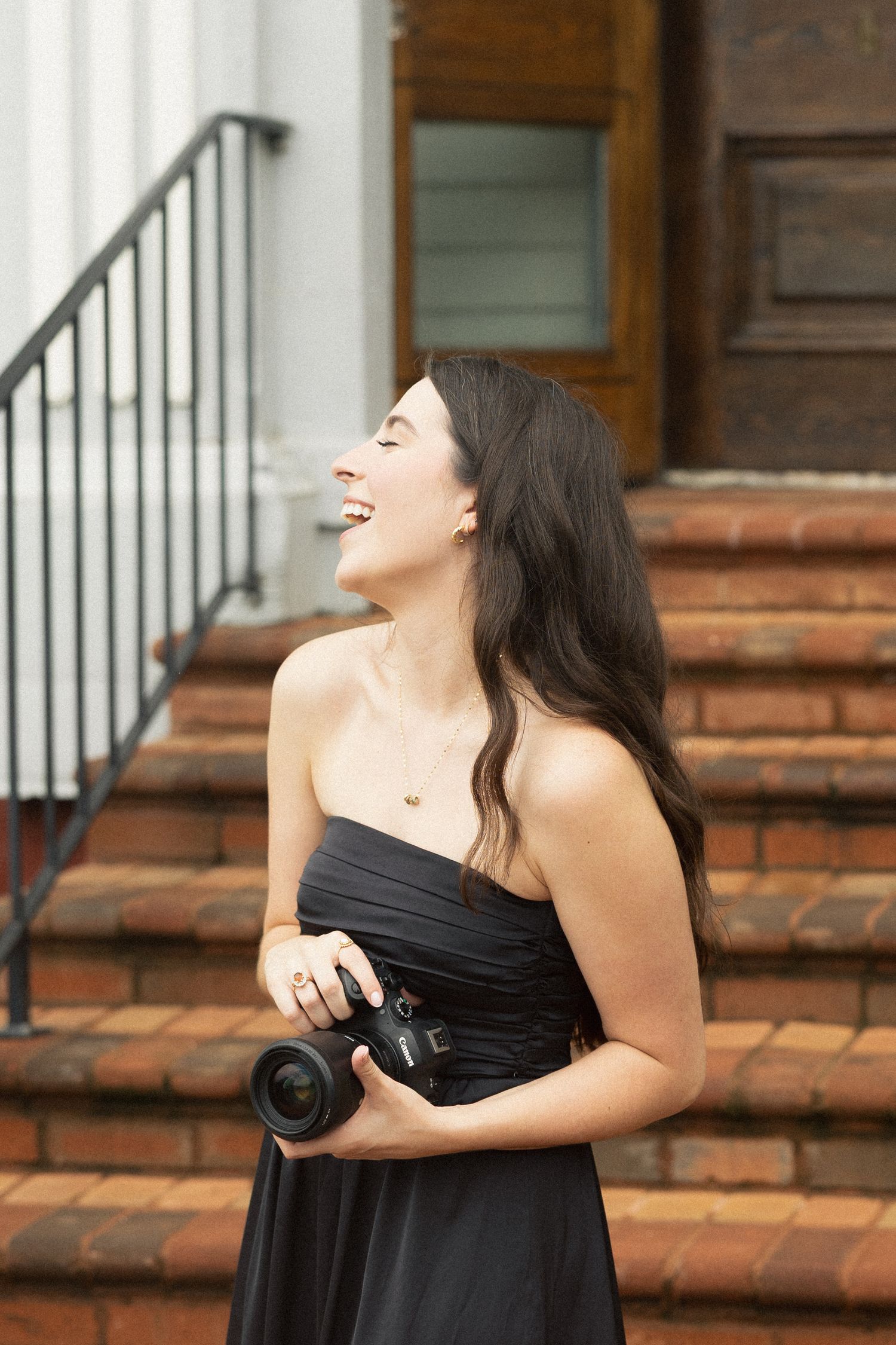 A laughing woman in a black strapless dress holds a DSLR camera while standing on brick steps outside a building.
