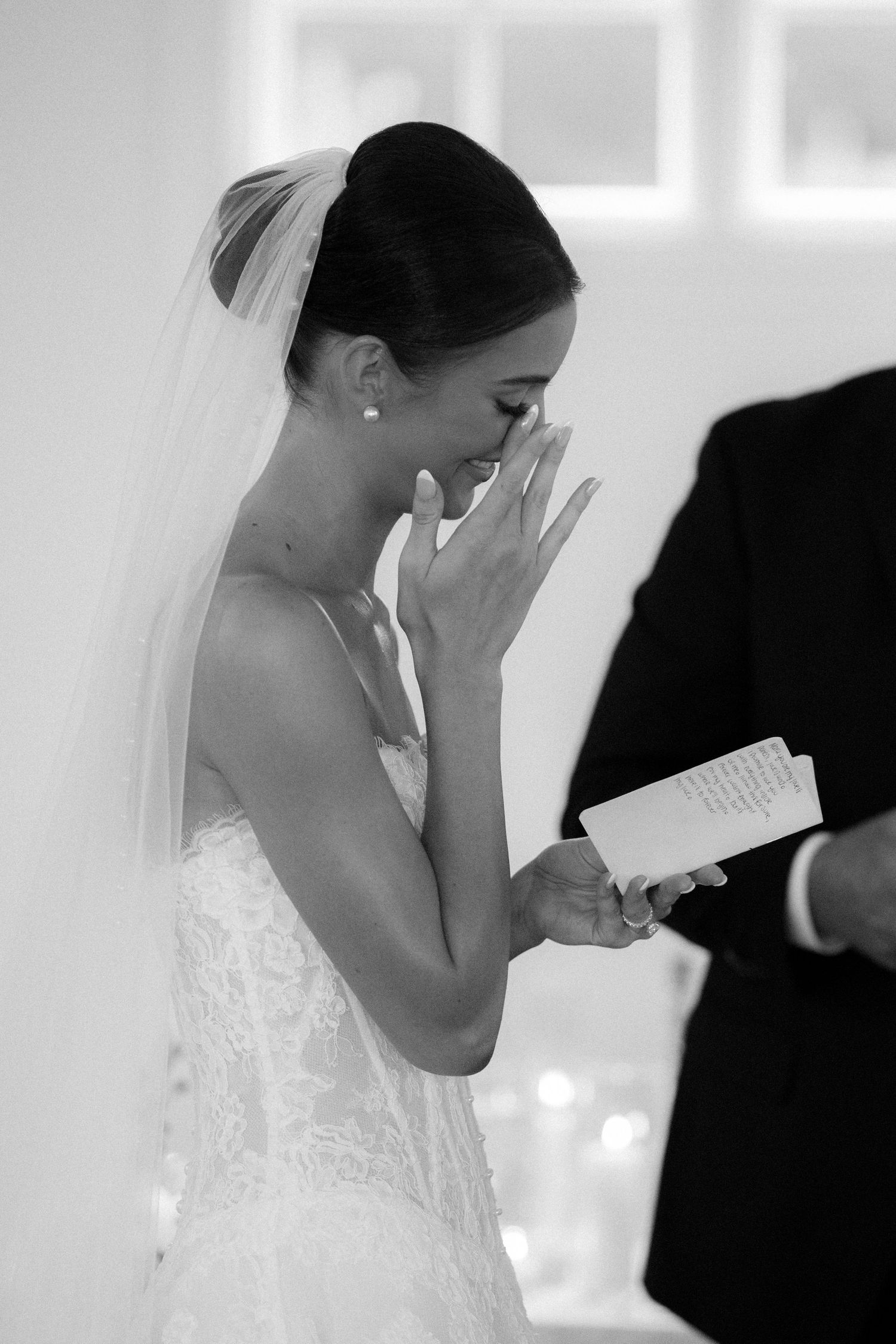 Emotional bride in lace wedding dress and veil wiping tears while reading vows, black and white photo.