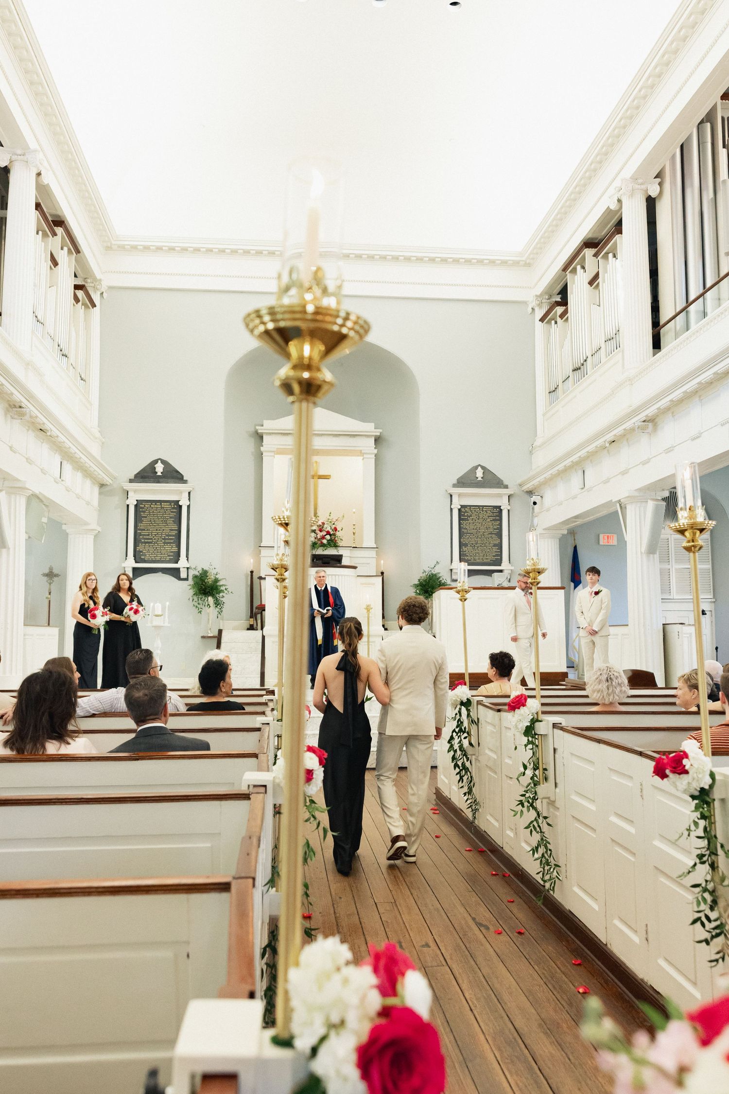 Wide view of wedding processional in elegant white church with pews adorned with floral garlands.
