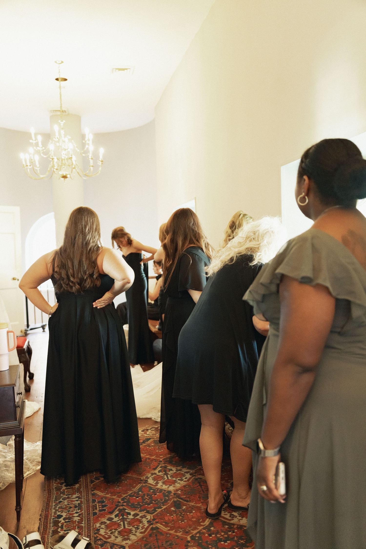 Bridesmaids in black gowns helping bride dress under chandelier in bright hallway.