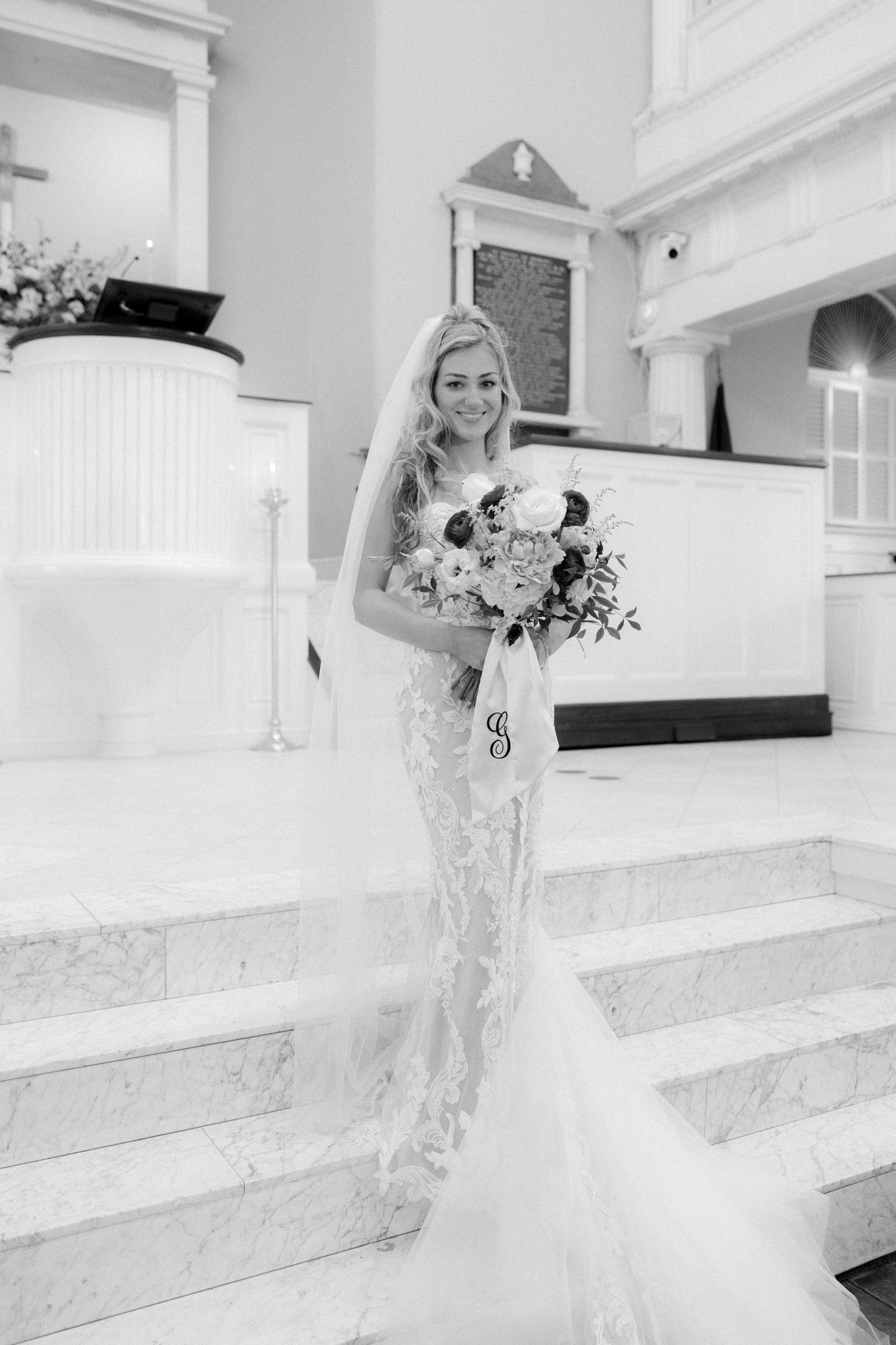 Black and white photo of bride in lace mermaid dress holding bouquet on marble church altar steps.