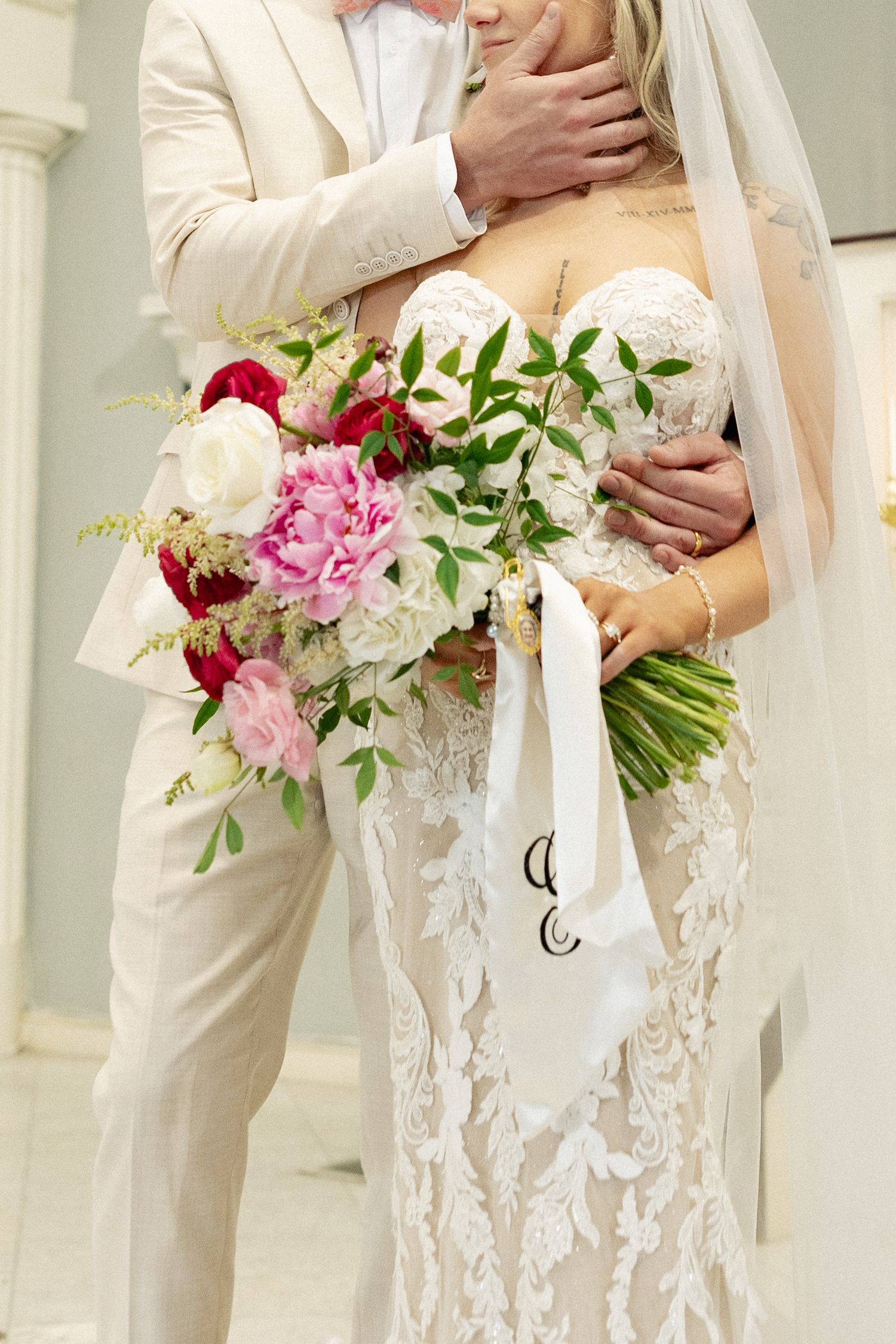 Bride in lace gown holding pink and red floral bouquet with white ribbon, embraced by groom in cream suit.