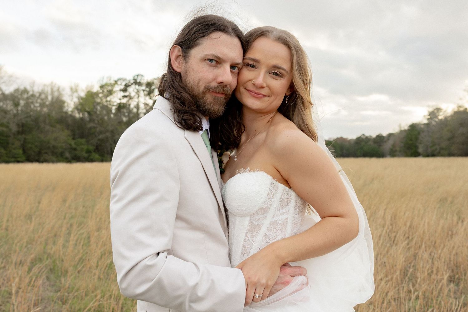 Smiling bride and groom embrace in a golden field with dramatic cloudy sky backdrop.