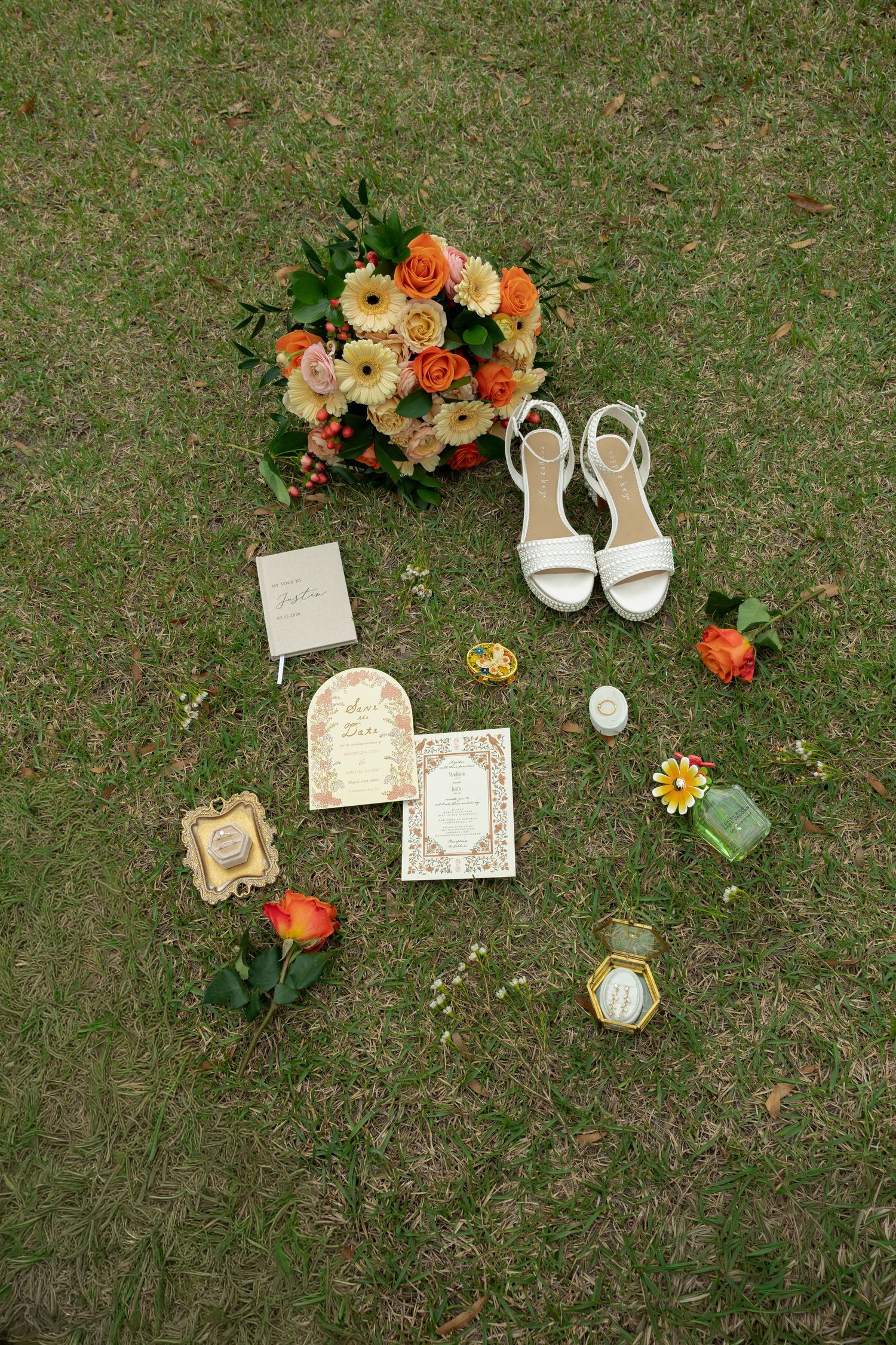 Elegant bridal flat lay with orange floral bouquet, white strappy heels, invitations, and gold accessories on grass.